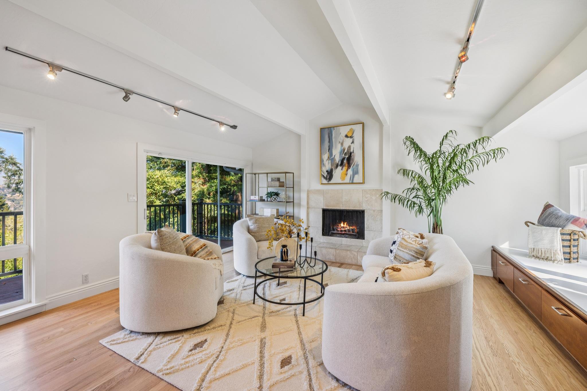 Bright living room with beige furniture, a fireplace, large windows showing trees outside, a ceiling track lighting system, a round glass coffee table with decorative objects, an abstract painting above the fireplace, a tall green potted plant, and a