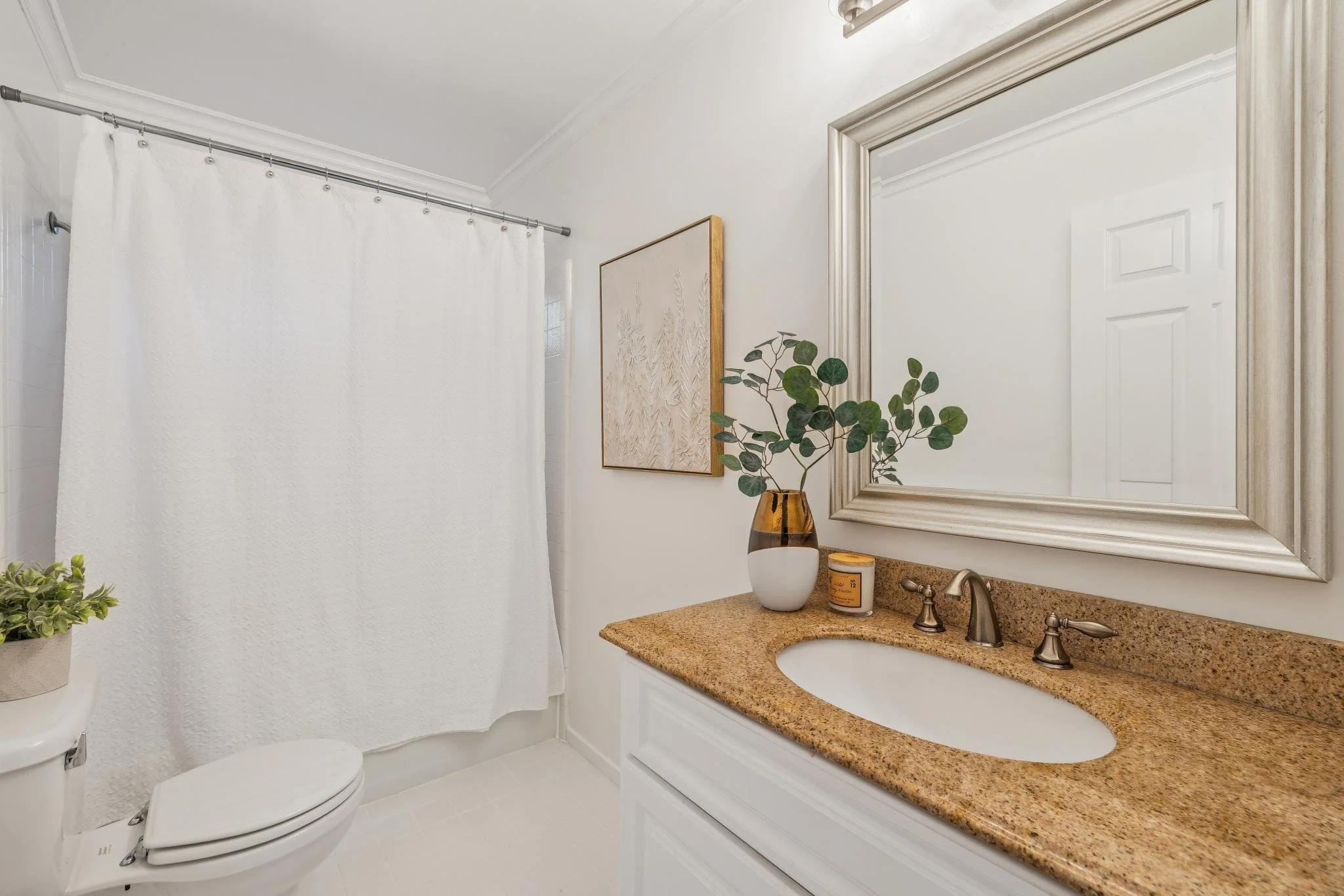Bathroom with white walls, a white toilet, a beige granite countertop with a sink, a large framed mirror, a decorative plant in a vase, a framed picture on the wall, a candle, and a white shower curtain on a silver rod.