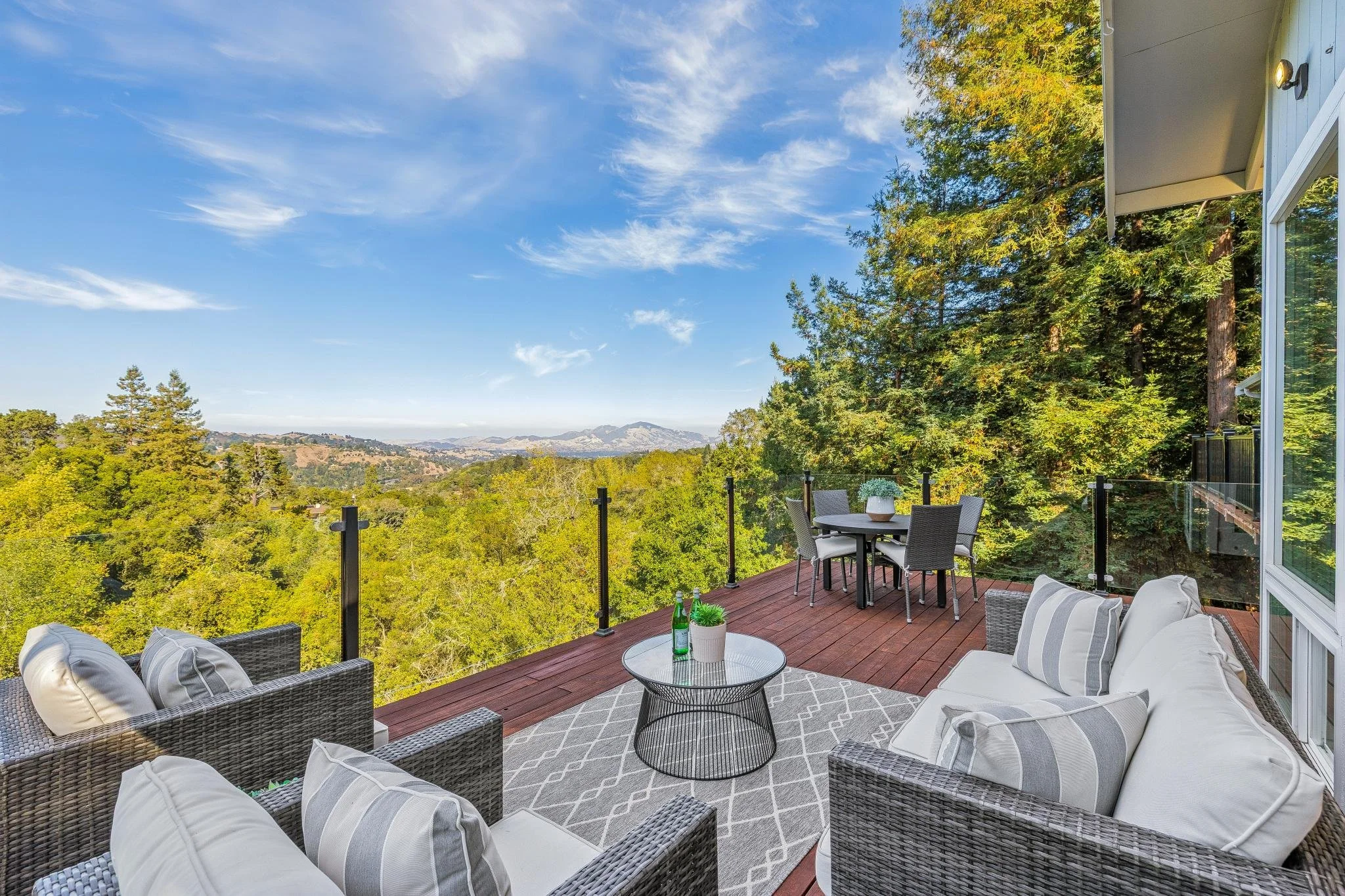 A spacious outdoor patio with outdoor furniture, including a dining table with chairs, a cushioned sofa, armchairs, and a coffee table with drinks. The patio has a glass railing, surrounded by lush green trees and overlooking a hilly landscape with d