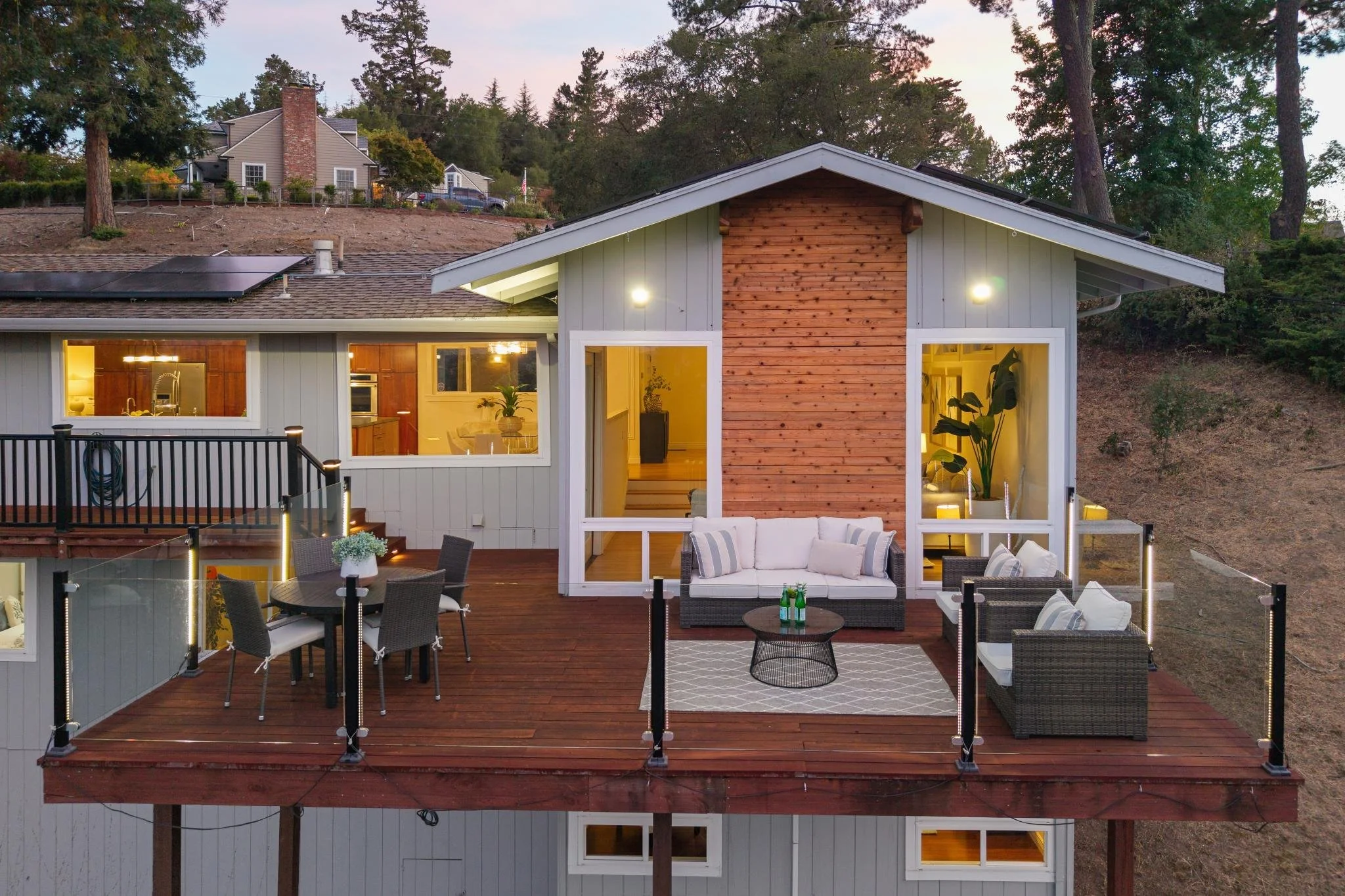 A house with a large wooden deck and outdoor seating area, illuminated at dusk, with a hillside and other houses in the background.