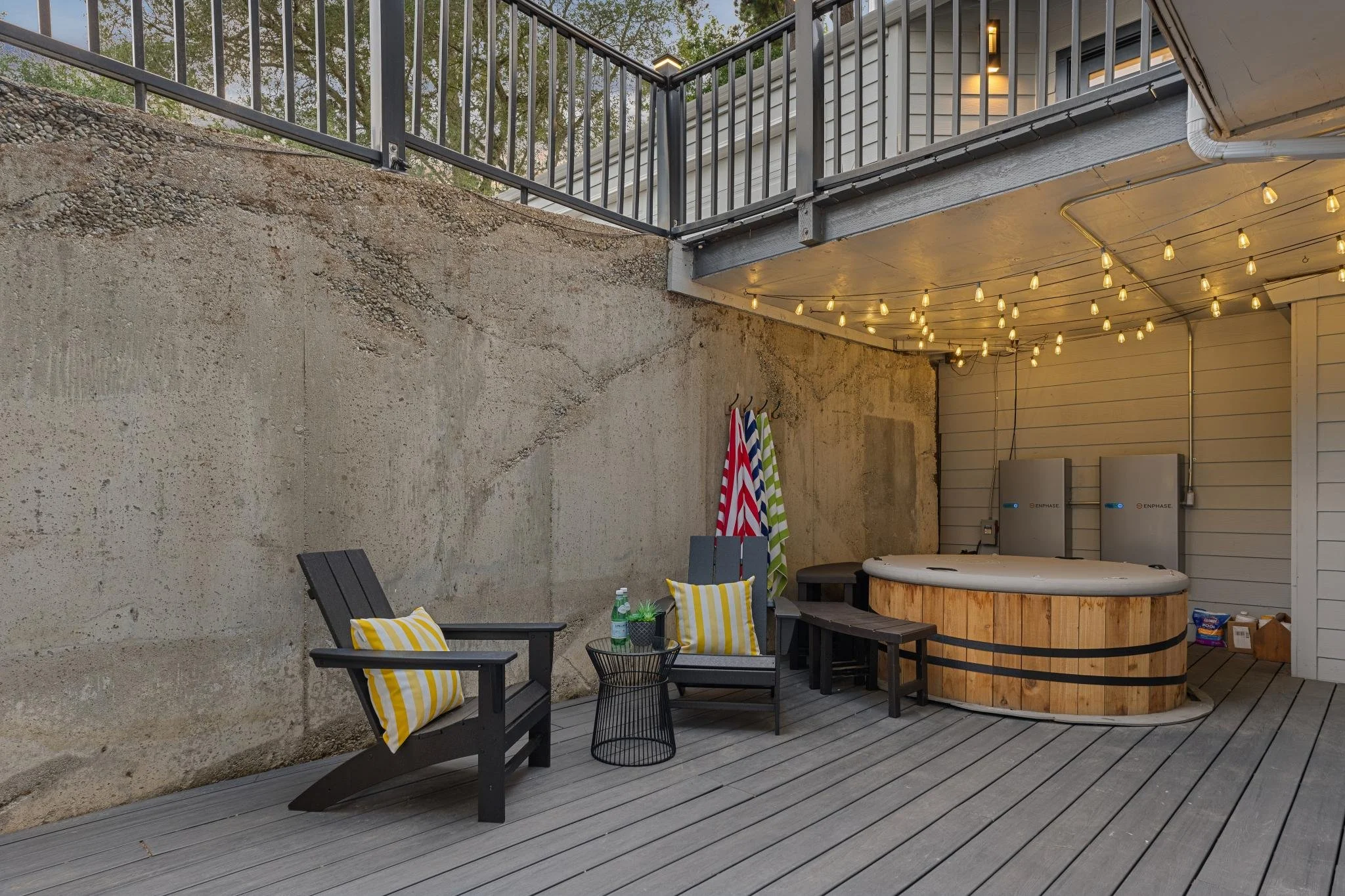 A backyard patio with black outdoor furniture, yellow striped cushions, and a hot tub. String lights are hanging under the deck, and there are towels hanging on hooks on the wall. The area has a wooden deck flooring and concrete wall.