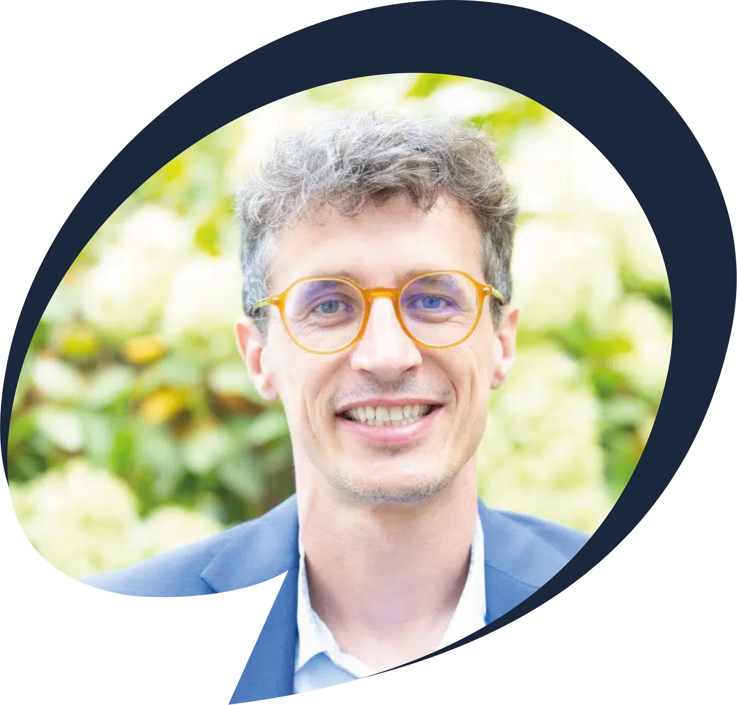 Close-up of our head of school, Jeremie, a smiling man with curly gray hair wearing orange glasses, blue suit, and white shirt, outdoors with blurred green foliage background.