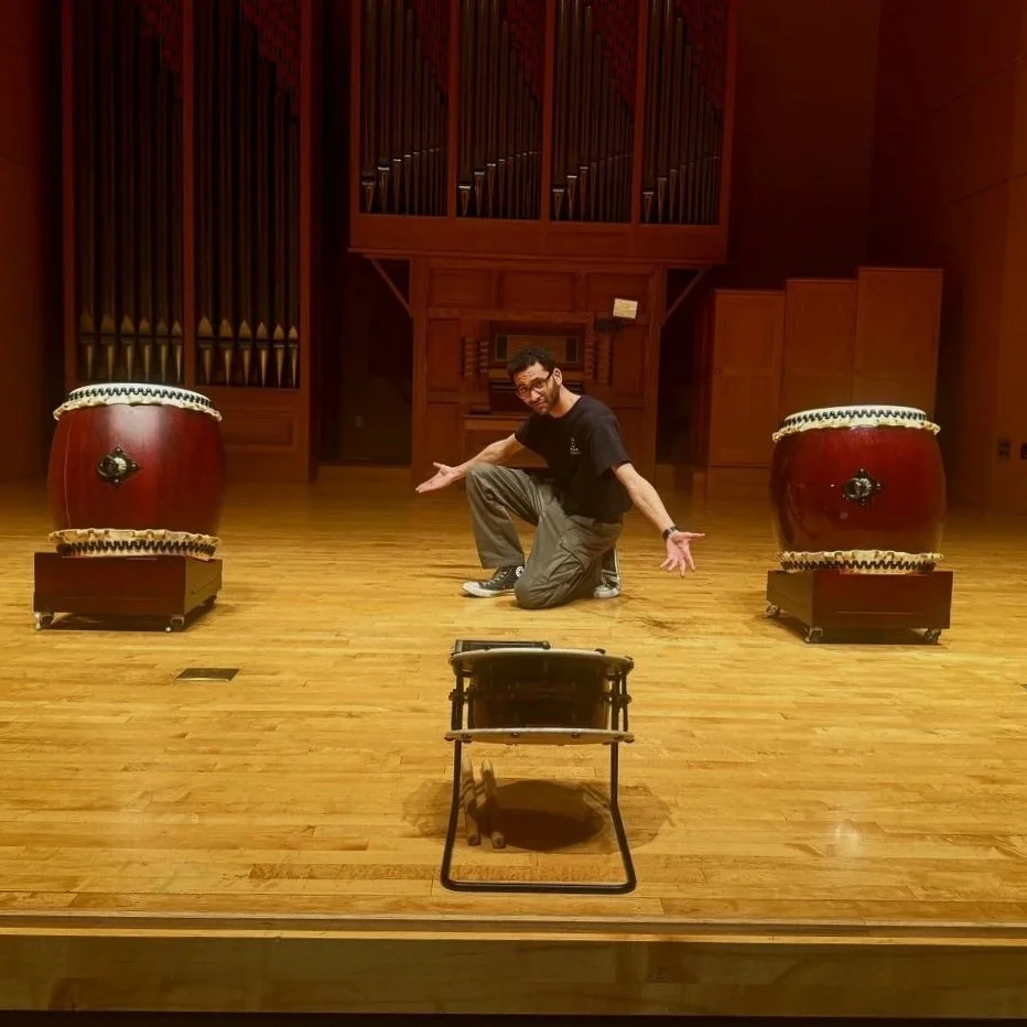 Man in black shirt and khaki pants kneeling on stage with arms outstretched, flanked by two large traditional drums. A smaller drum is in front of him on the stage floor, in a concert hall with wooden floors and wall panels.
