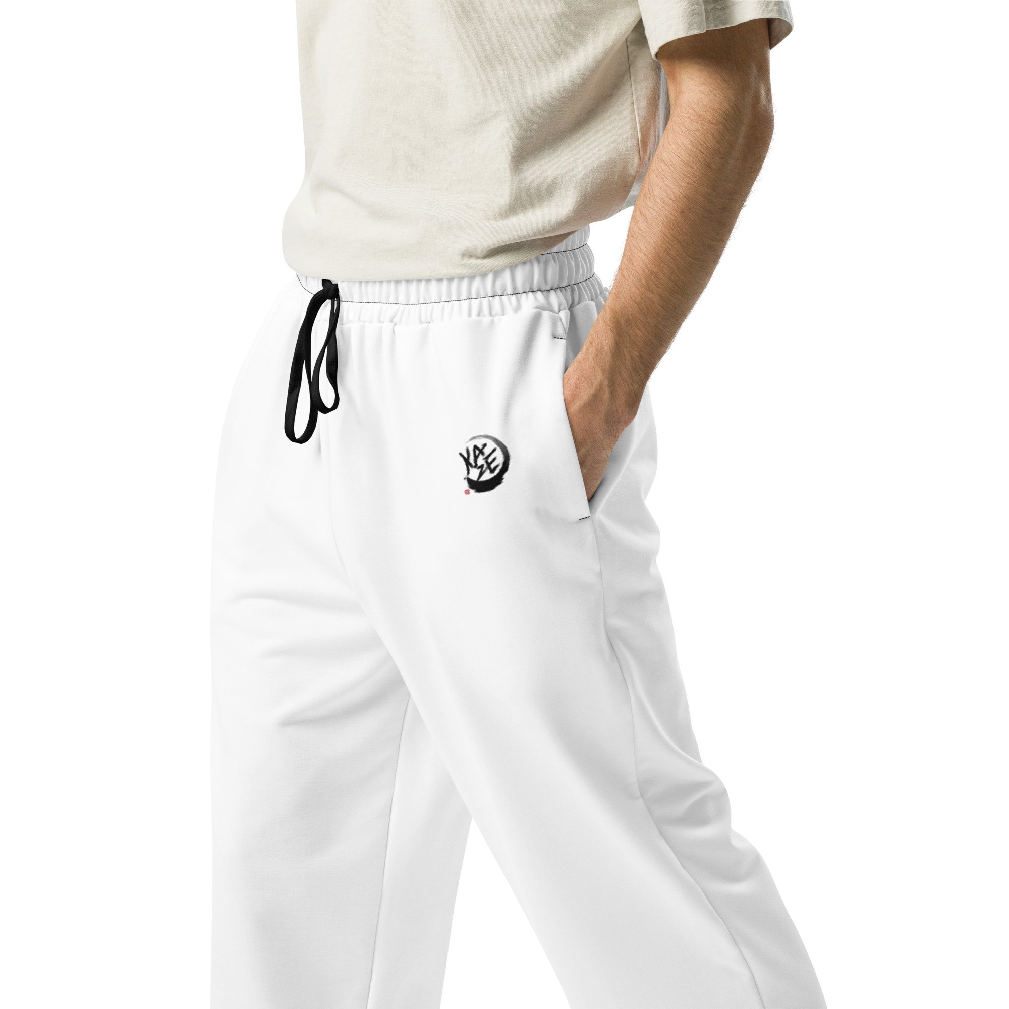 Person wearing white athletic pants with a black drawstring and a logo, and a beige T-shirt, with one hand in the pocket.