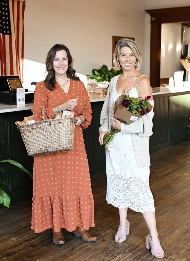 Two women standing inside a room, one holding a basket of groceries and the other holding a bouquet of flowers, in front of a counter with plants and a cash register.
