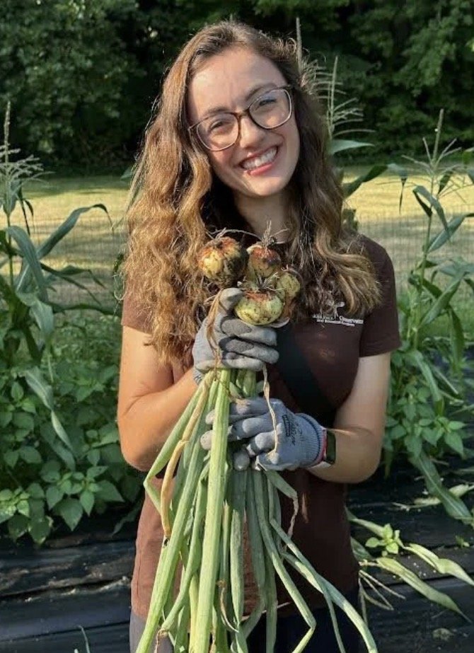 Young woman with glasses and wavy hair holding freshly harvested onions in a garden.