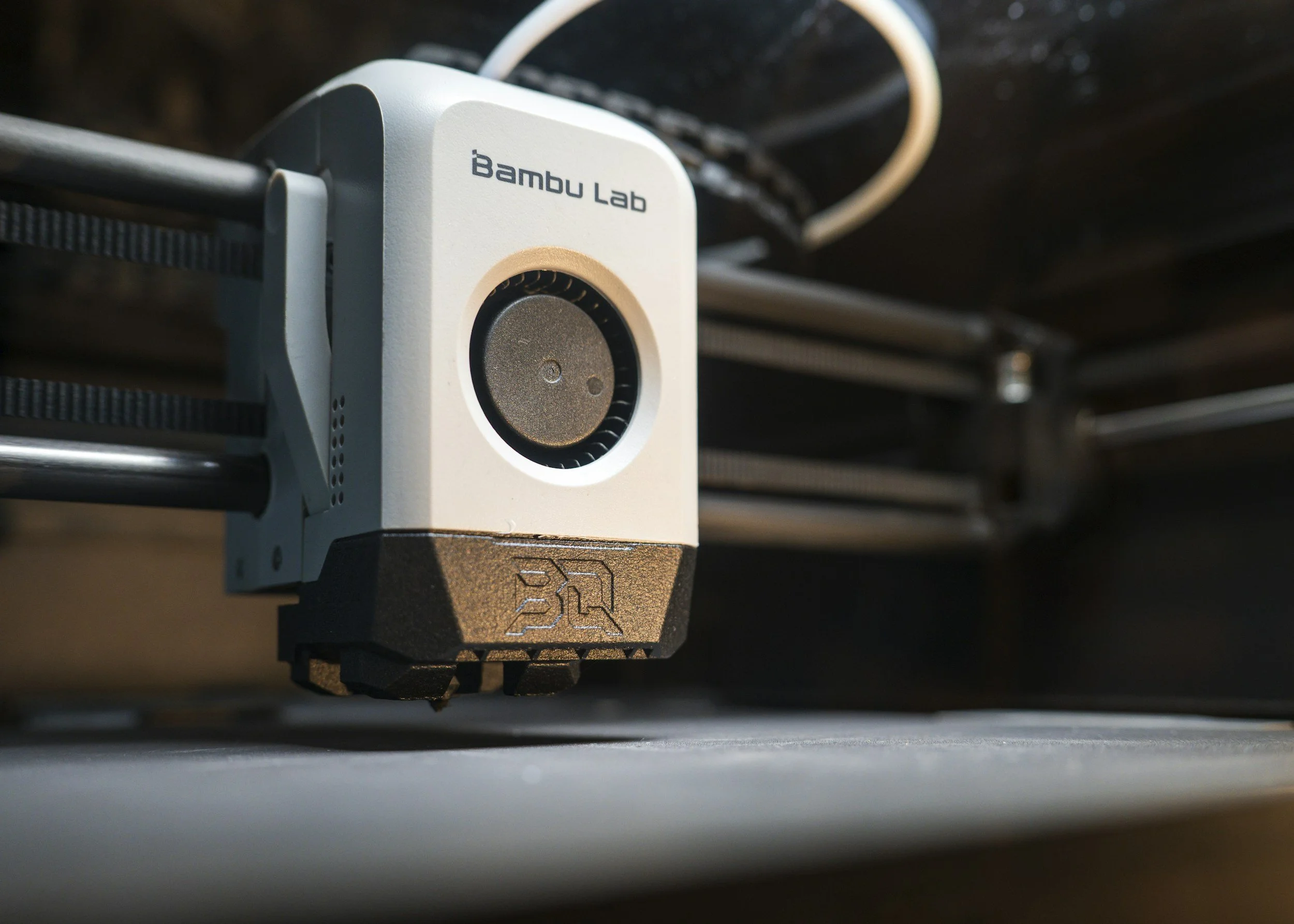 Close-up of a white 3D printer extruder with the品牌 name 'Bambu Lab' printed on it, moving along a print bed with metal rods and black frame components visible in the background.
