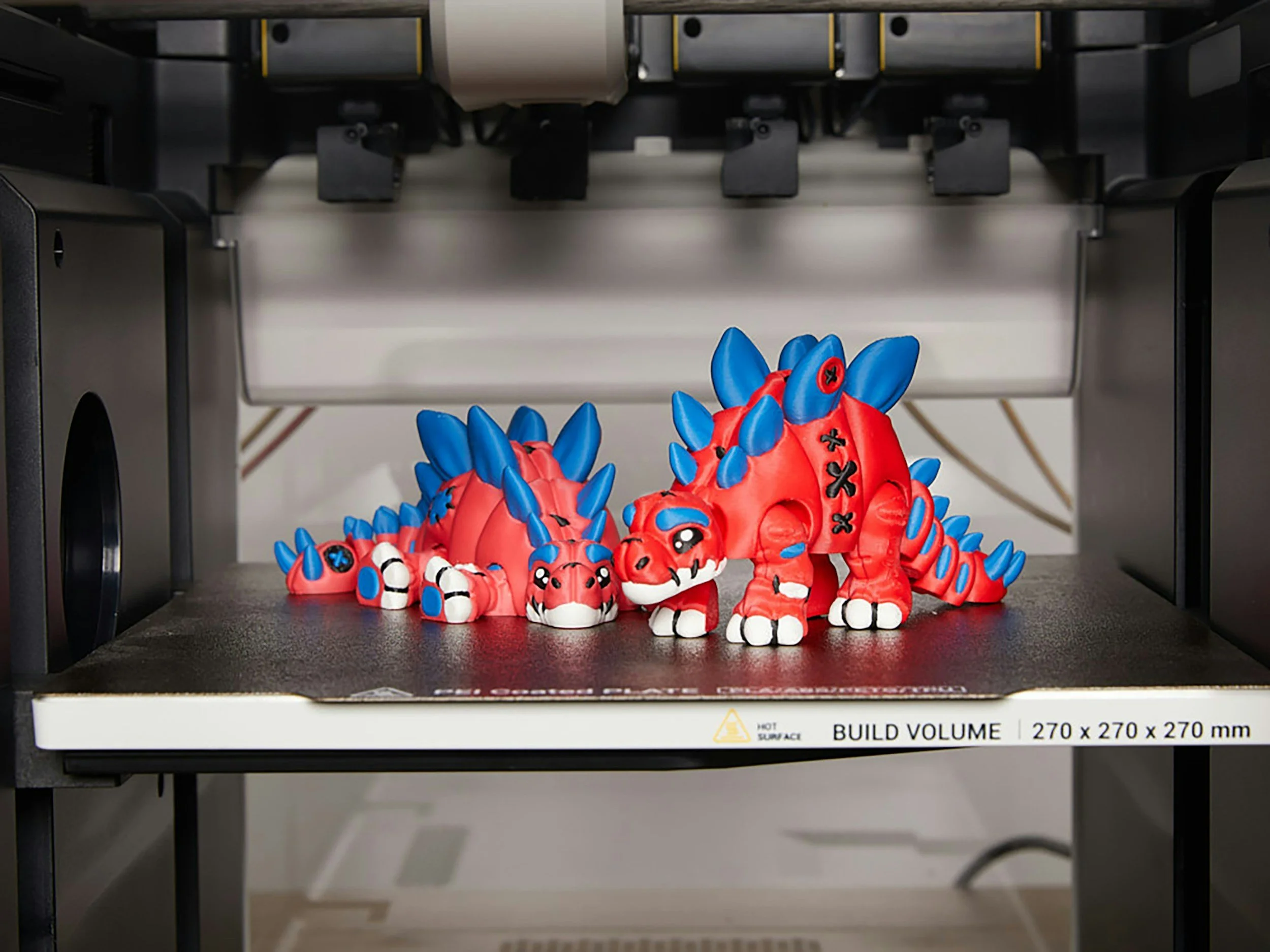 3D-printed red and blue stylized dinosaur toys on a 3D printer build platform.