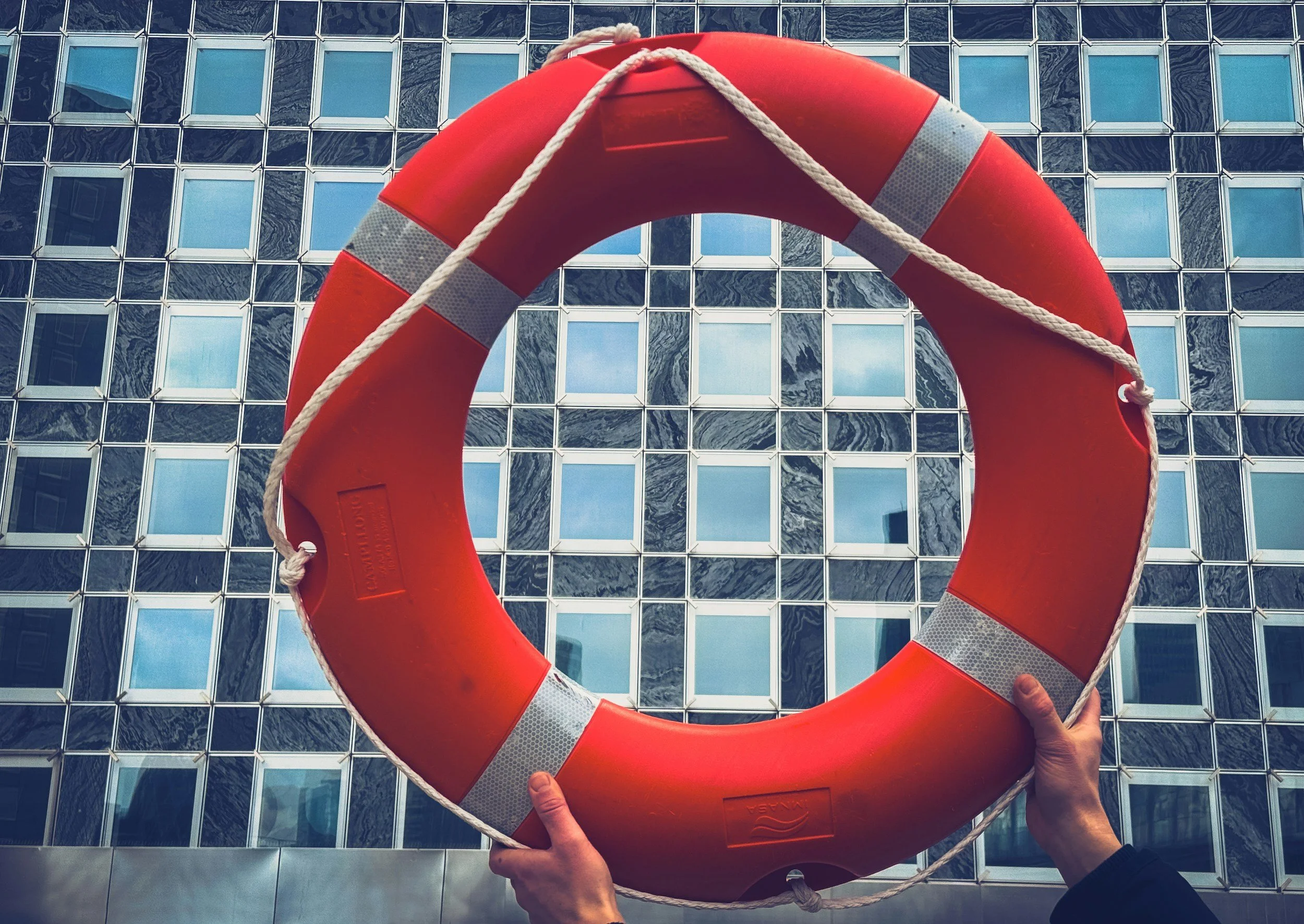 A person holding a red lifebuoy with white reflective strips in front of a building with a grid of reflective blue windows.