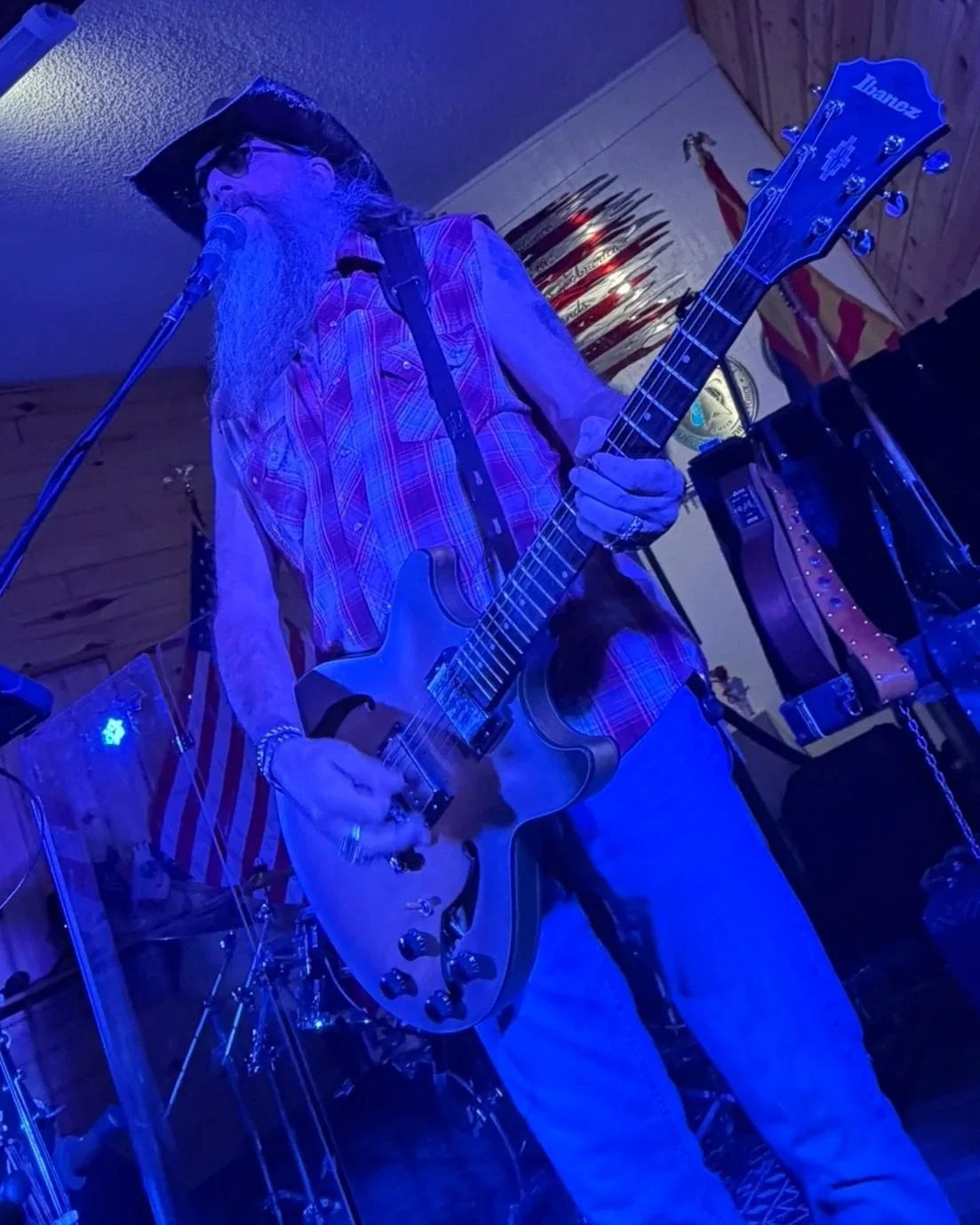 A bearded man wearing a cowboy hat, plaid shirt, and jeans playing an electric guitar on stage under blue lighting inside a wooden-paneled venue decorated with flags, including an American flag.