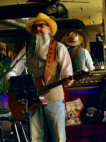 A man with a beard, wearing sunglasses, a yellow hat, a beige vest, and blue jeans, is playing an acoustic guitar and singing into a microphone in a cozy indoor setting with musical instruments and decorations.
