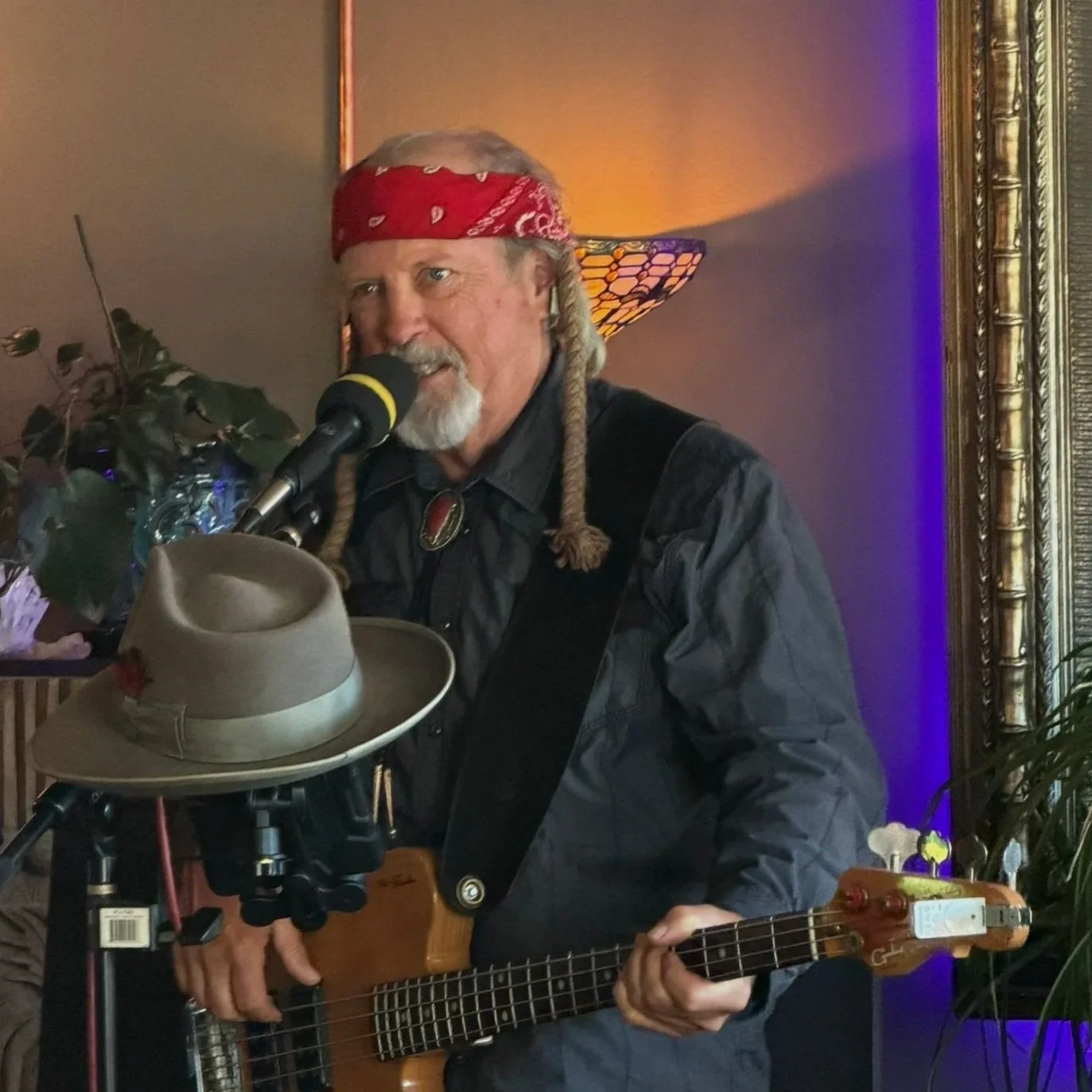 A man with a red bandana and gray beard playing an acoustic guitar and singing into a microphone at an indoor event.