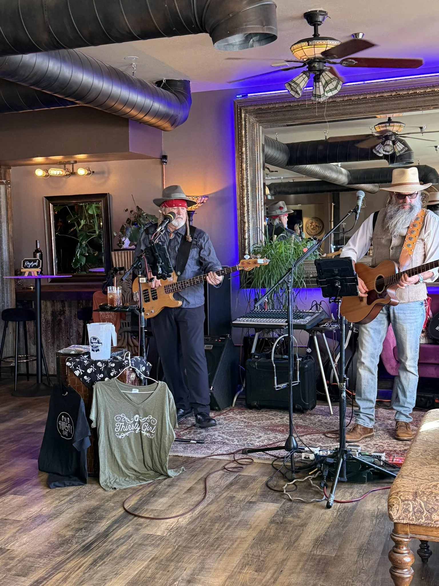 Two musicians performing with guitars, one wearing a cowboy hat and red bandana, the other wearing a wide-brimmed straw hat with sunglasses. They are in a cozy bar or cafe with a large mirror, plants, and decorative lighting, and there is sound equipment and merchandise on display.