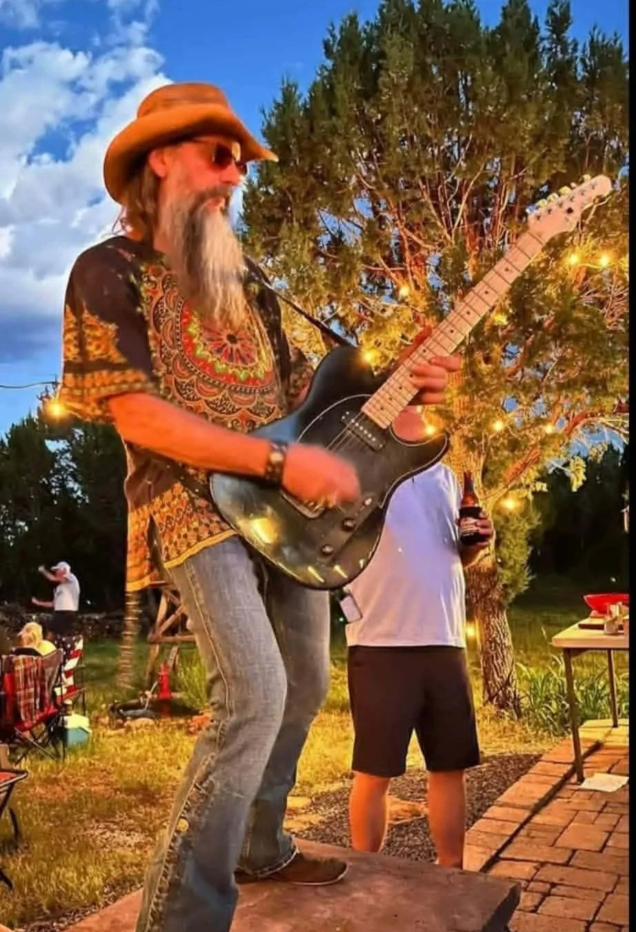 A man with long hair and a beard wearing a cowboy hat, sunglasses, and a colorful patterned shirt is playing an electric guitar outdoors during evening time. Behind him, there is a tree with string lights, and in the background, a group of people gathered for a casual outdoor event.