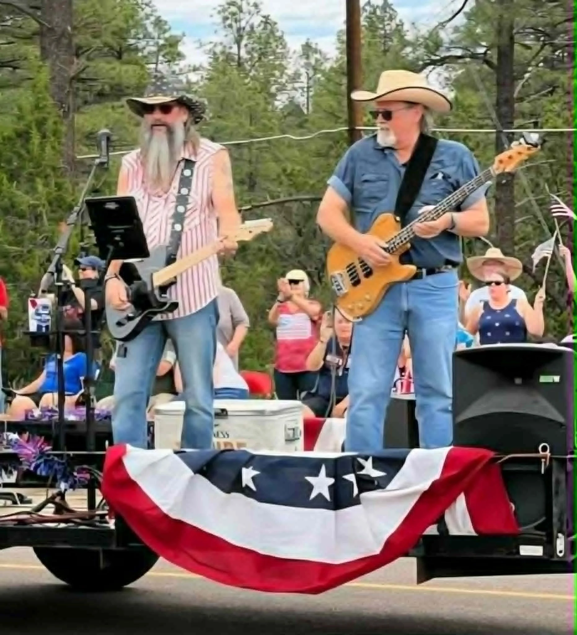 Two men wearing cowboy hats and sunglasses standing on a float decorated with American flag-themed fabric during a parade. One man is playing a guitar, and the other has a beard and striped sleeveless shirt. Spectators are watching in the background.