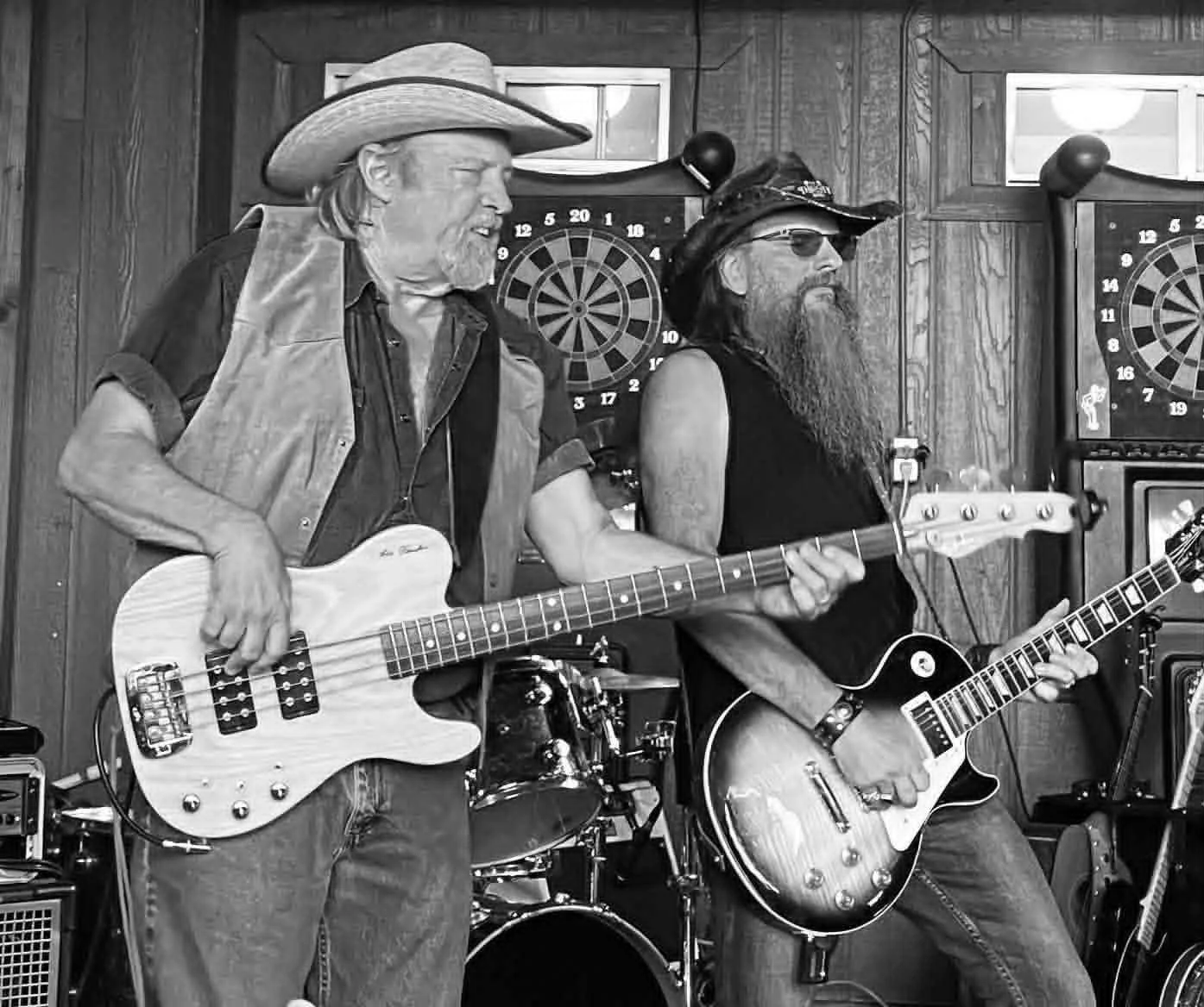 Two men with long hair and beards playing electric guitars and a bass guitar in a wooden-paneled room with dartboards on the wall, suggesting a casual musical jam session.