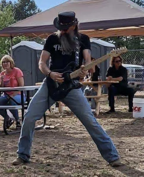 A man wearing a cowboy hat and sunglasses playing an electric guitar outdoors during a gathering or event.