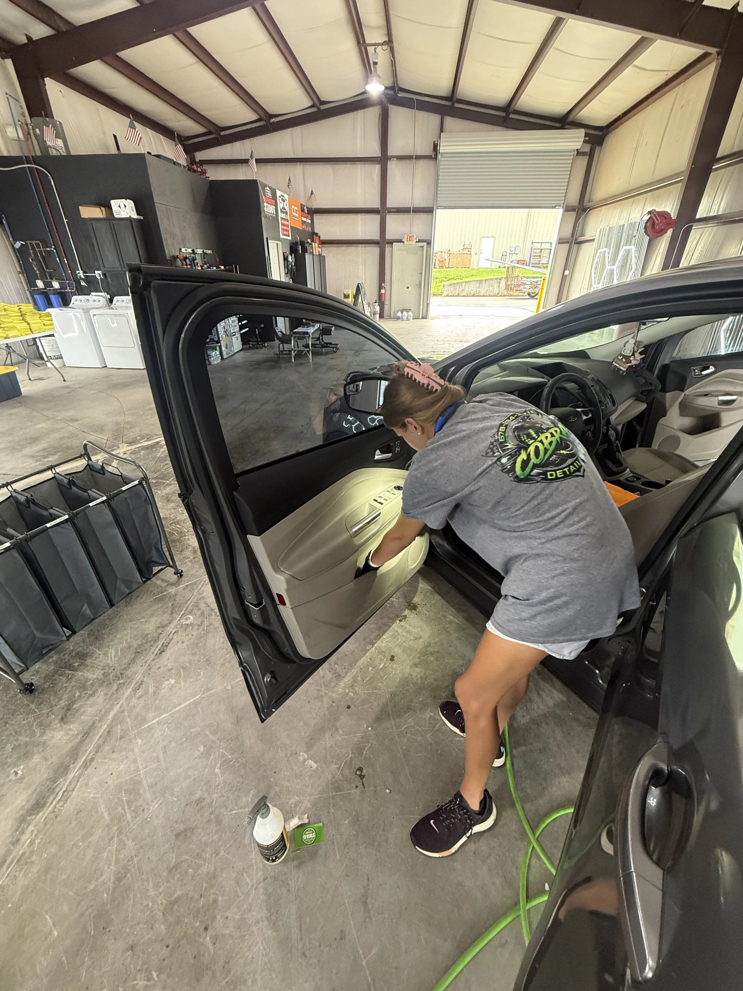 A person using a vacuum cleaner to clean the interior of a black car inside a spacious garage with a high ceiling, several chairs, and various tools and supplies.