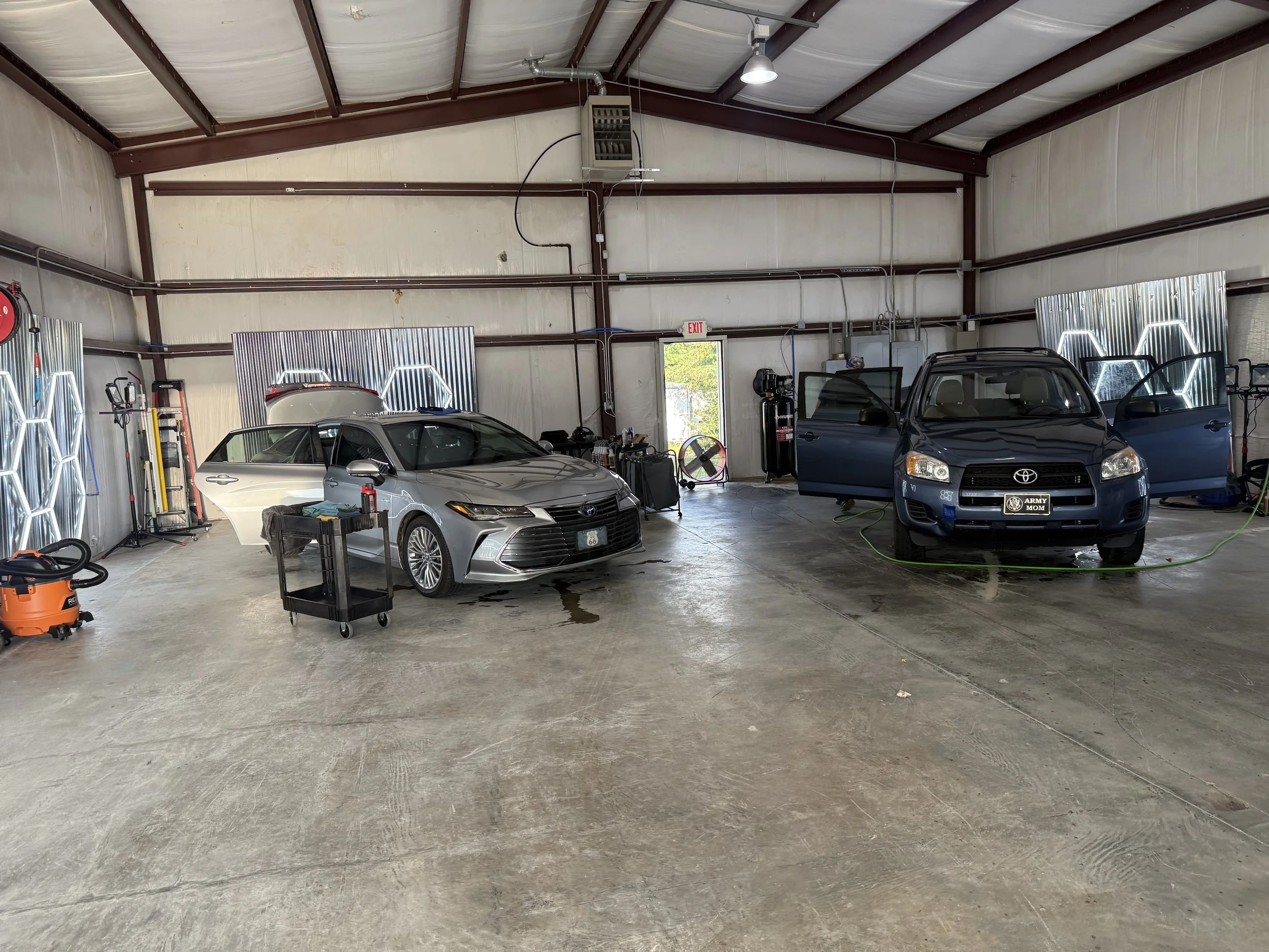 Indoor garage with two parked cars, tools and equipment, metal sheet walls, and industrial ceiling lights.