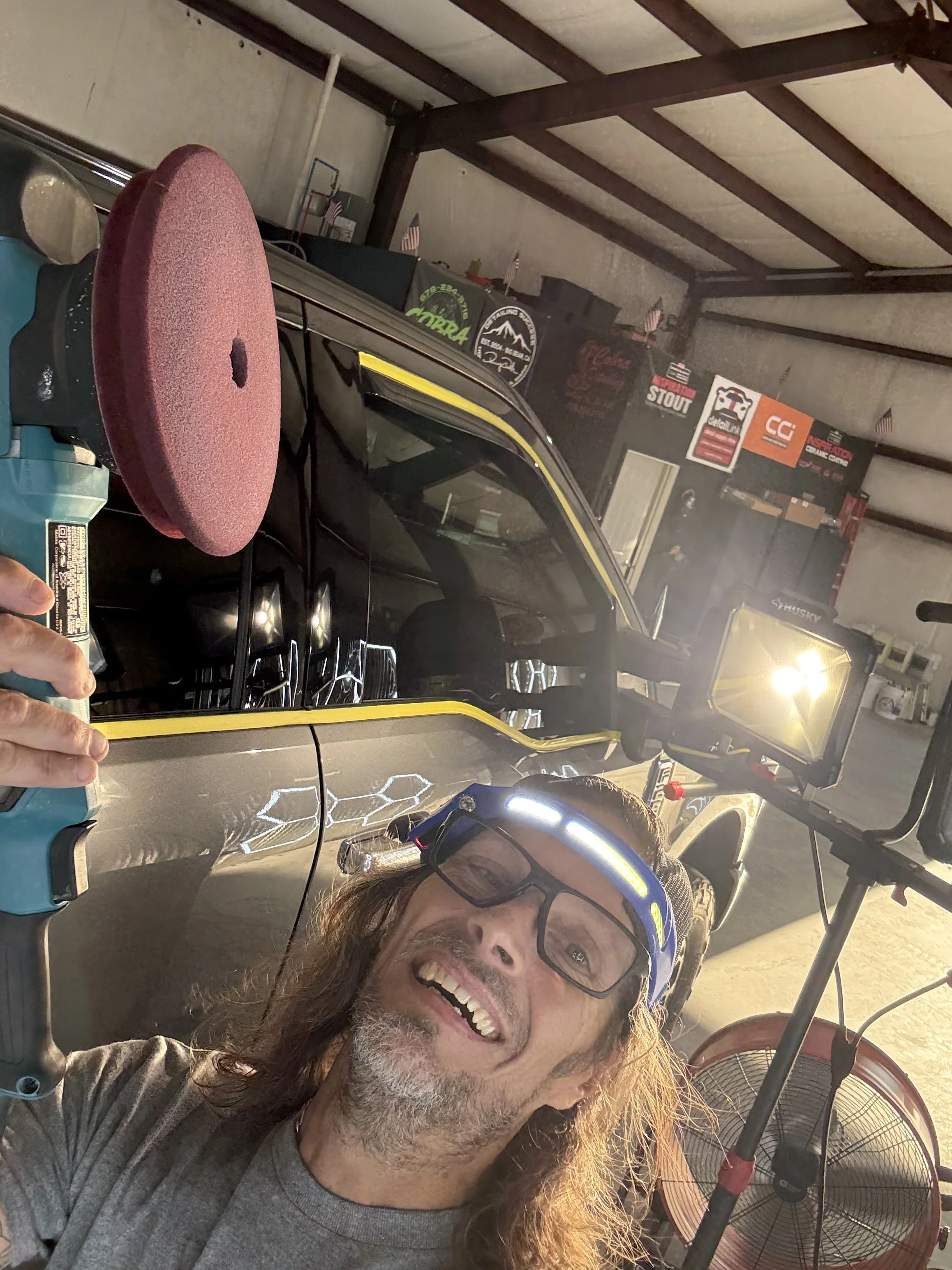 A man with long hair and glasses, smiling and wearing a gray shirt, taking a selfie inside a garage with various signs on the wall, a fan, and riding equipment including a golf cart.