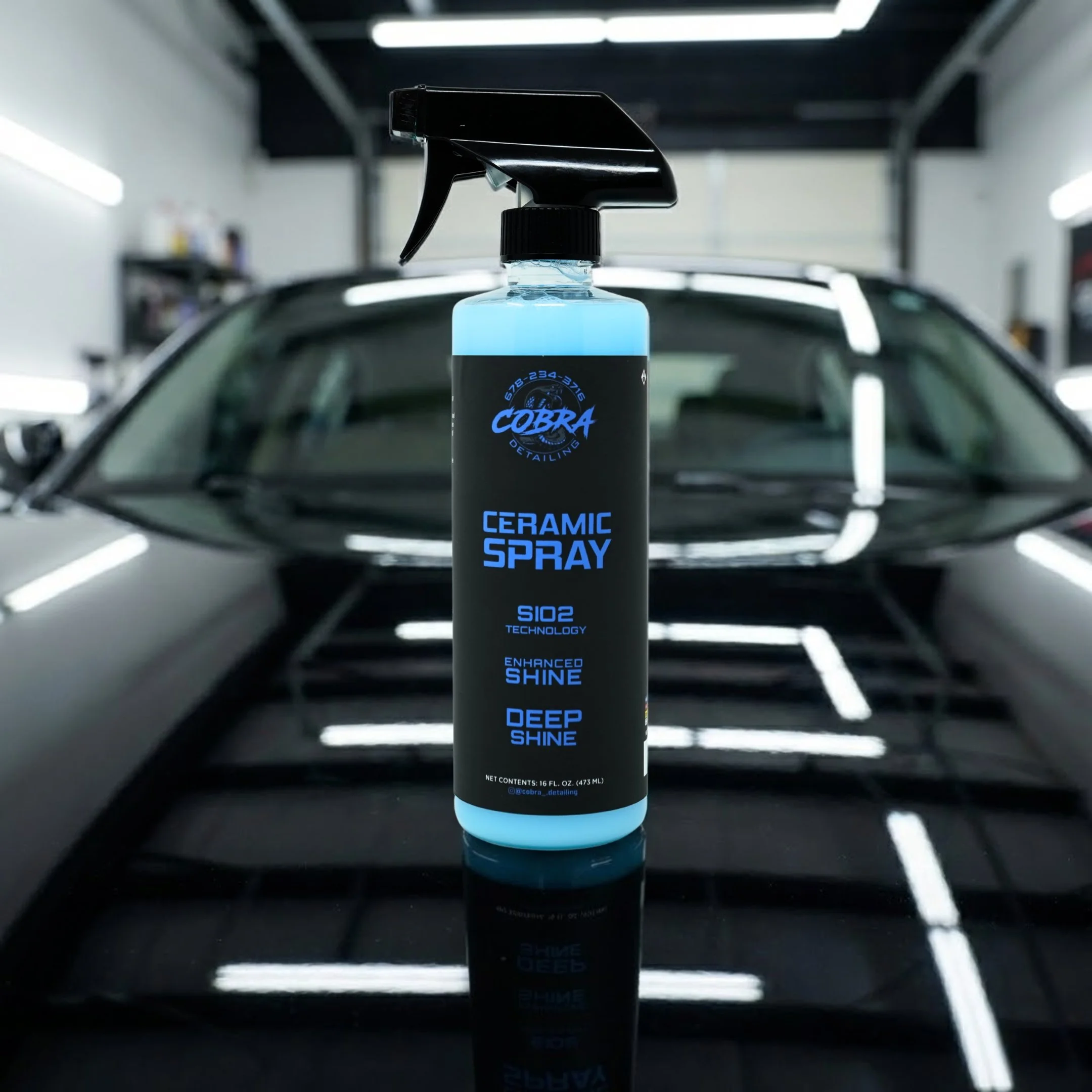 A spray bottle labeled 'Cobra Ceramic Spray' is placed on the hood of a black car in a garage or detailing shop with bright overhead lighting.