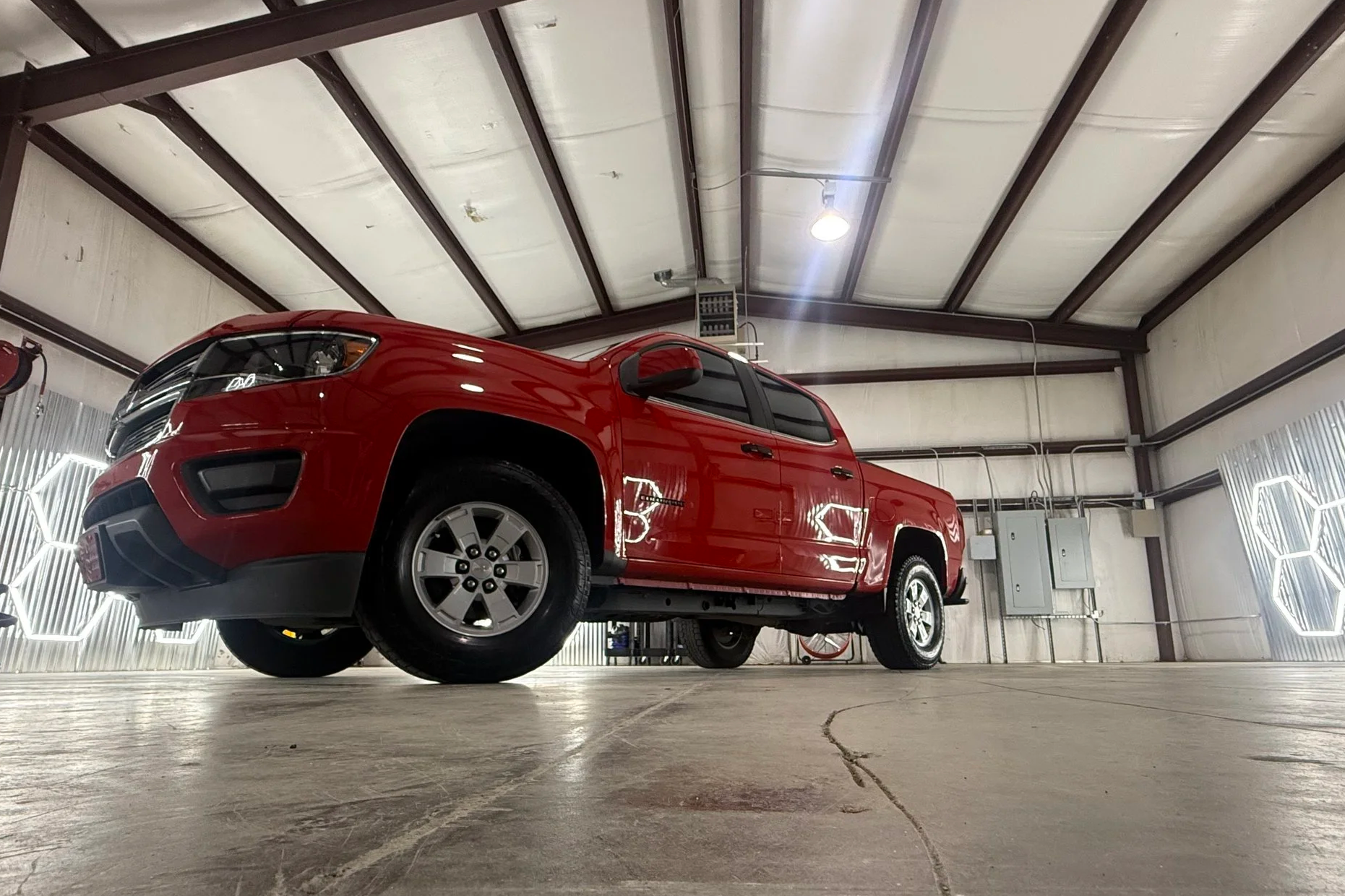 Red pickup truck parked inside a spacious garage with a high ceiling and exposed metal beams, illuminated by ceiling lights and decorative hexagonal light fixtures.