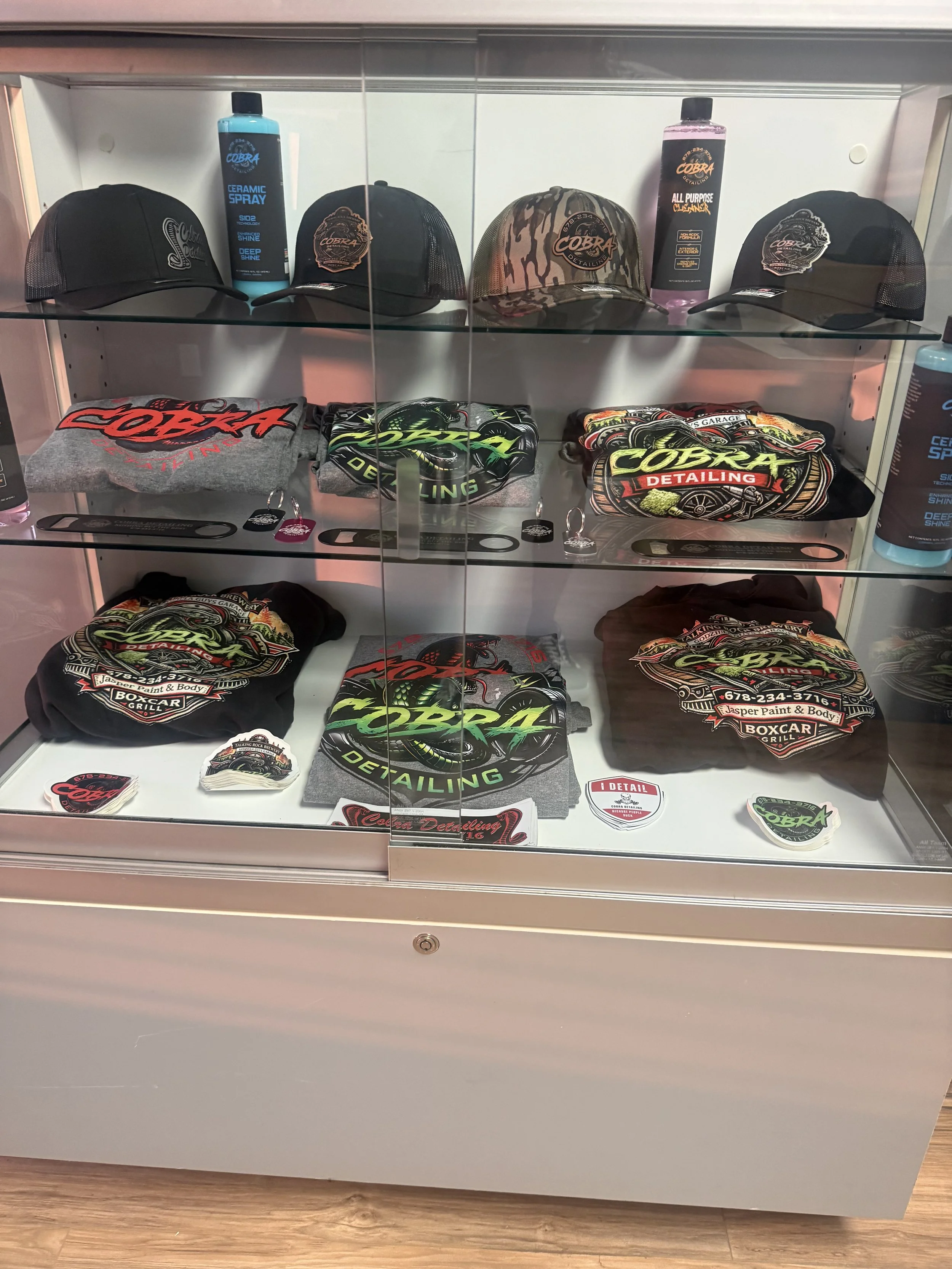 A display case with Cobra Detailing merchandise including baseball caps, t-shirts, and stickers. The items feature Cobra Detailing logos and graphics with bold text and vibrant colors.