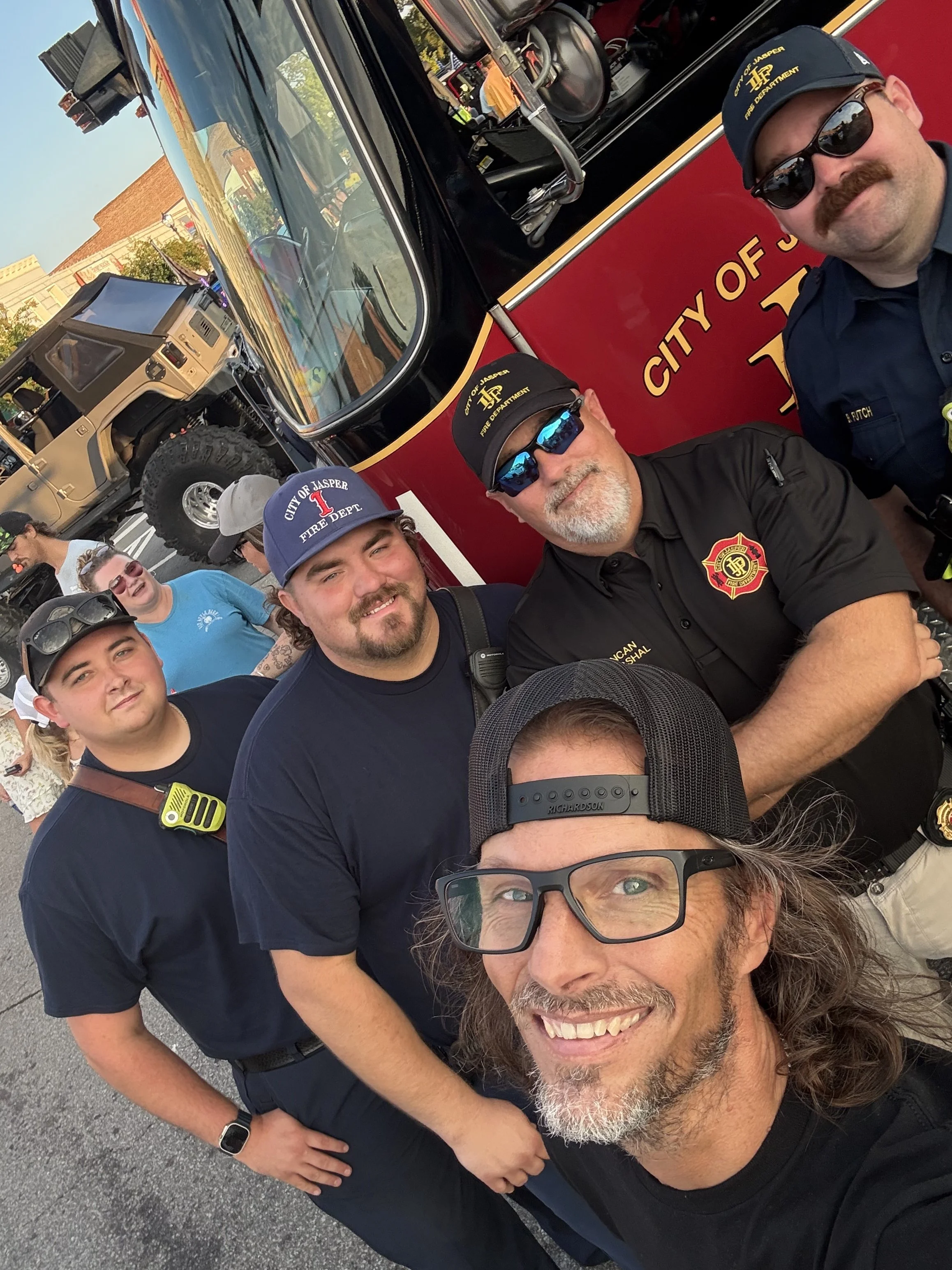A group of four men taking a selfie in front of a red fire truck. The men are wearing casual clothing and sunglasses, and appear to be firefighters or fire department personnel from the City of Jasper. The background shows a parking lot and some buildings.