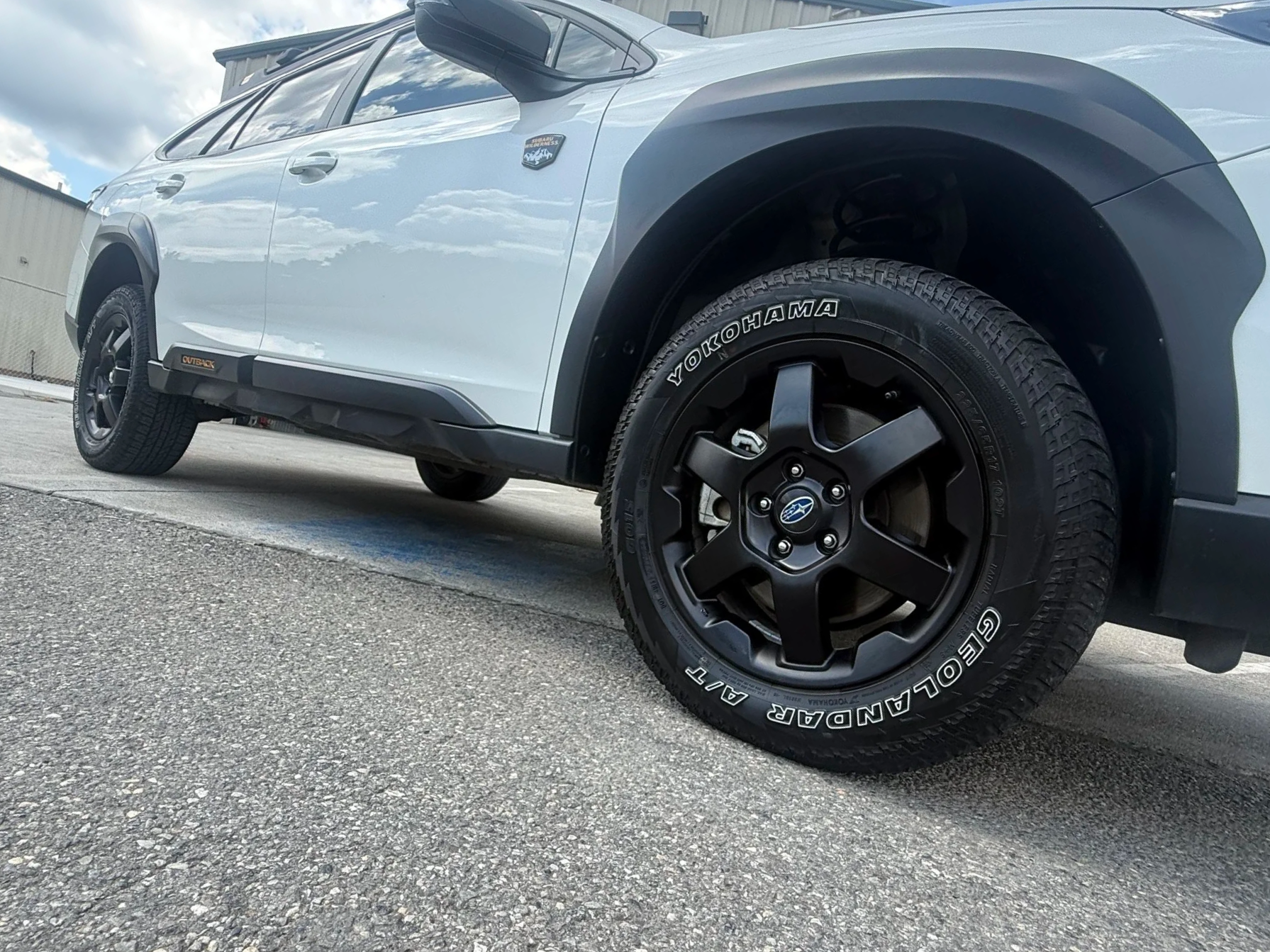 A white Subaru SUV with black wheels and Yokohama tires parked on a paved surface, with an overcast sky reflected in the windows.