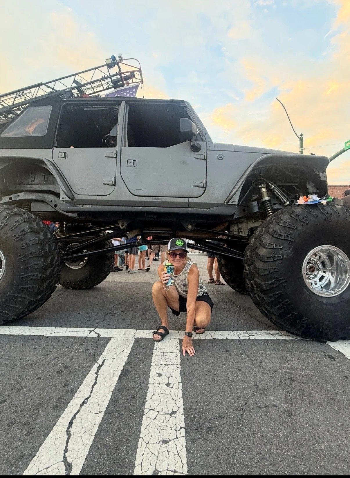A woman crouching under a large off-road vehicle with oversized tires, holding a drink and smiling at the camera, at an outdoor event during sunset with a crowd in the background.