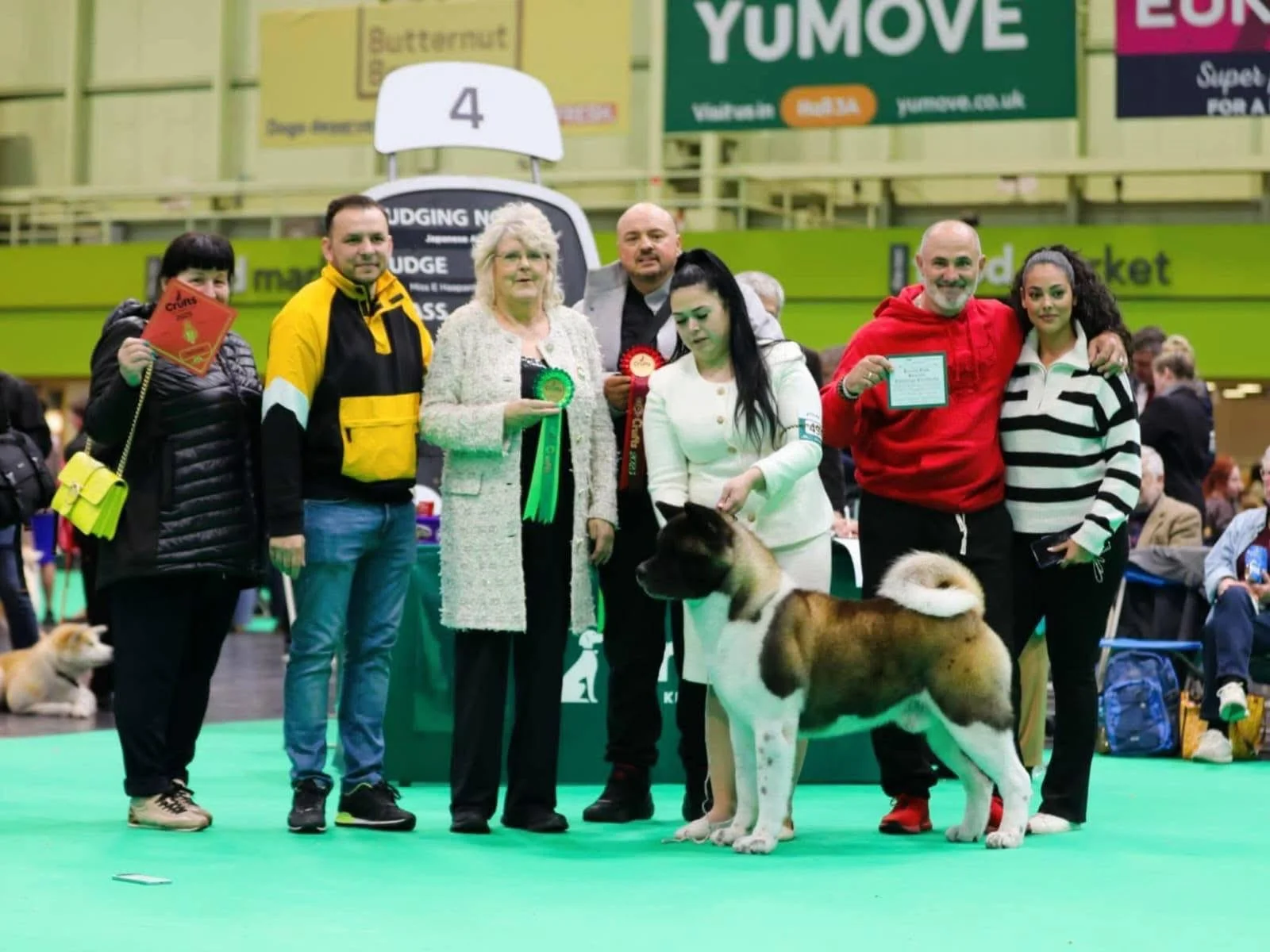 Group of people and dogs at a dog show, with some holding ribbons and awards, inside an indoor arena.