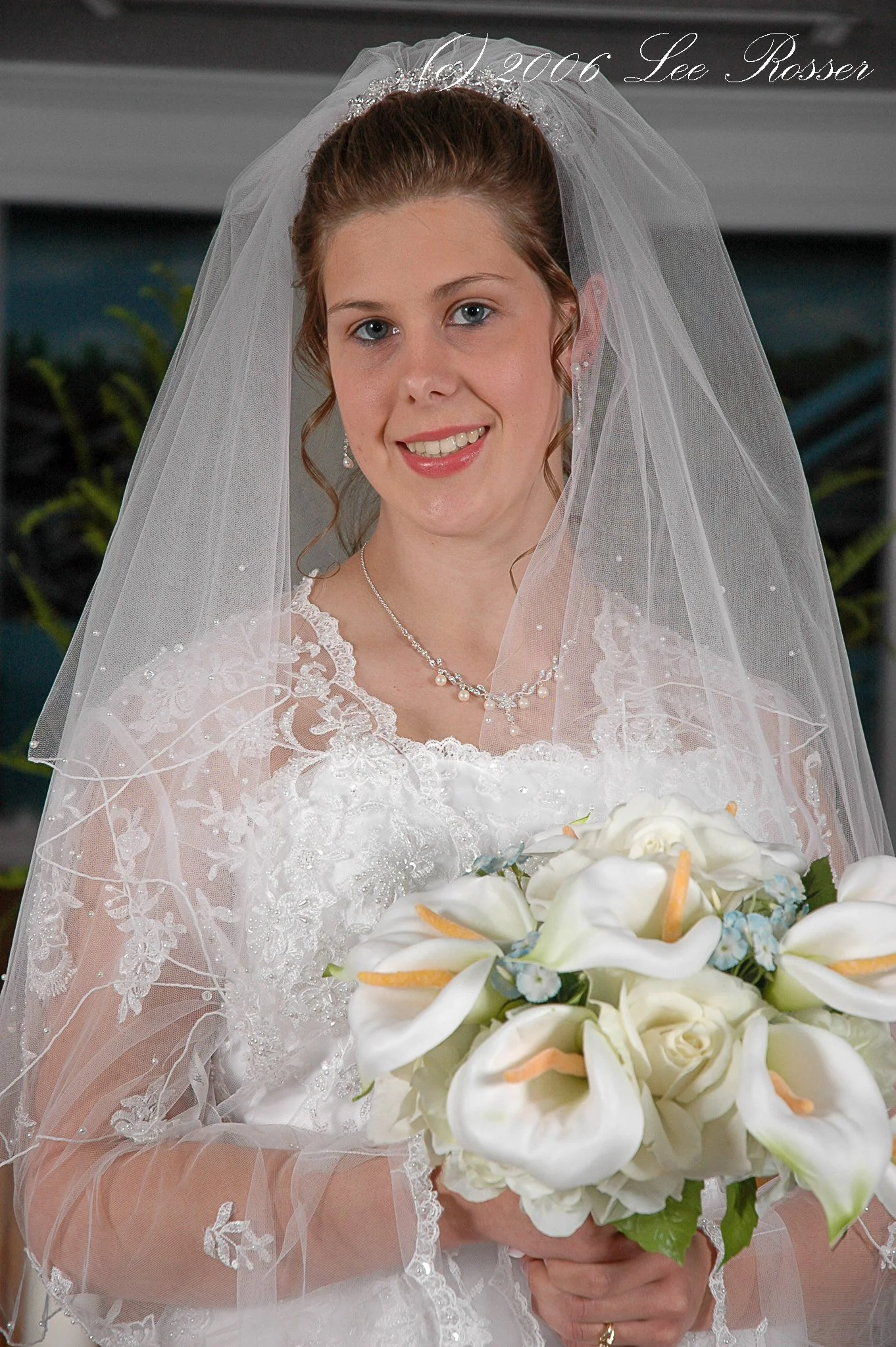 A bride with red hair styled in an updo, wearing a white lace wedding dress and a silver necklace, holding a bouquet of white calla lilies with some orange and blue accents, smiling with blue eyes. She is wearing a veil with delicate embroidery and a