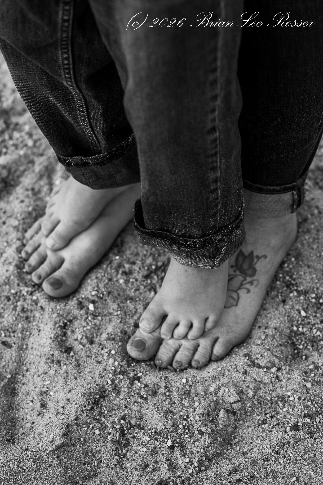 Close-up of a person's bare feet in the sand, with toes slightly spread and toenails painted. The person is wearing dark rolled-up jeans and a portion of a tattoo on one ankle is visible.