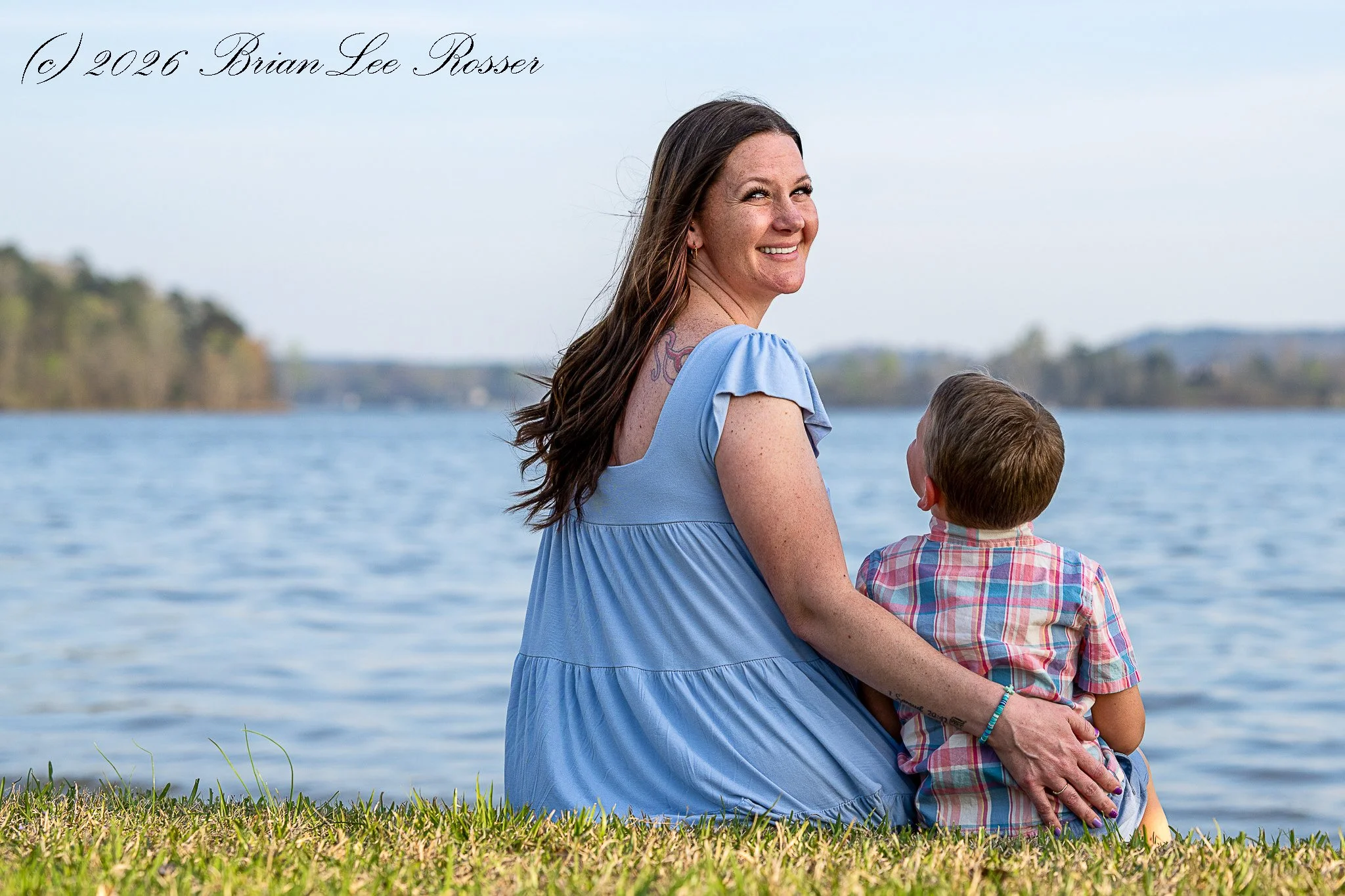 A woman and a young boy sit on the grass near a body of water, looking towards the water with a backdrop of distant trees and hills. The woman is smiling and has long brown hair, while the boy wears a plaid shirt.