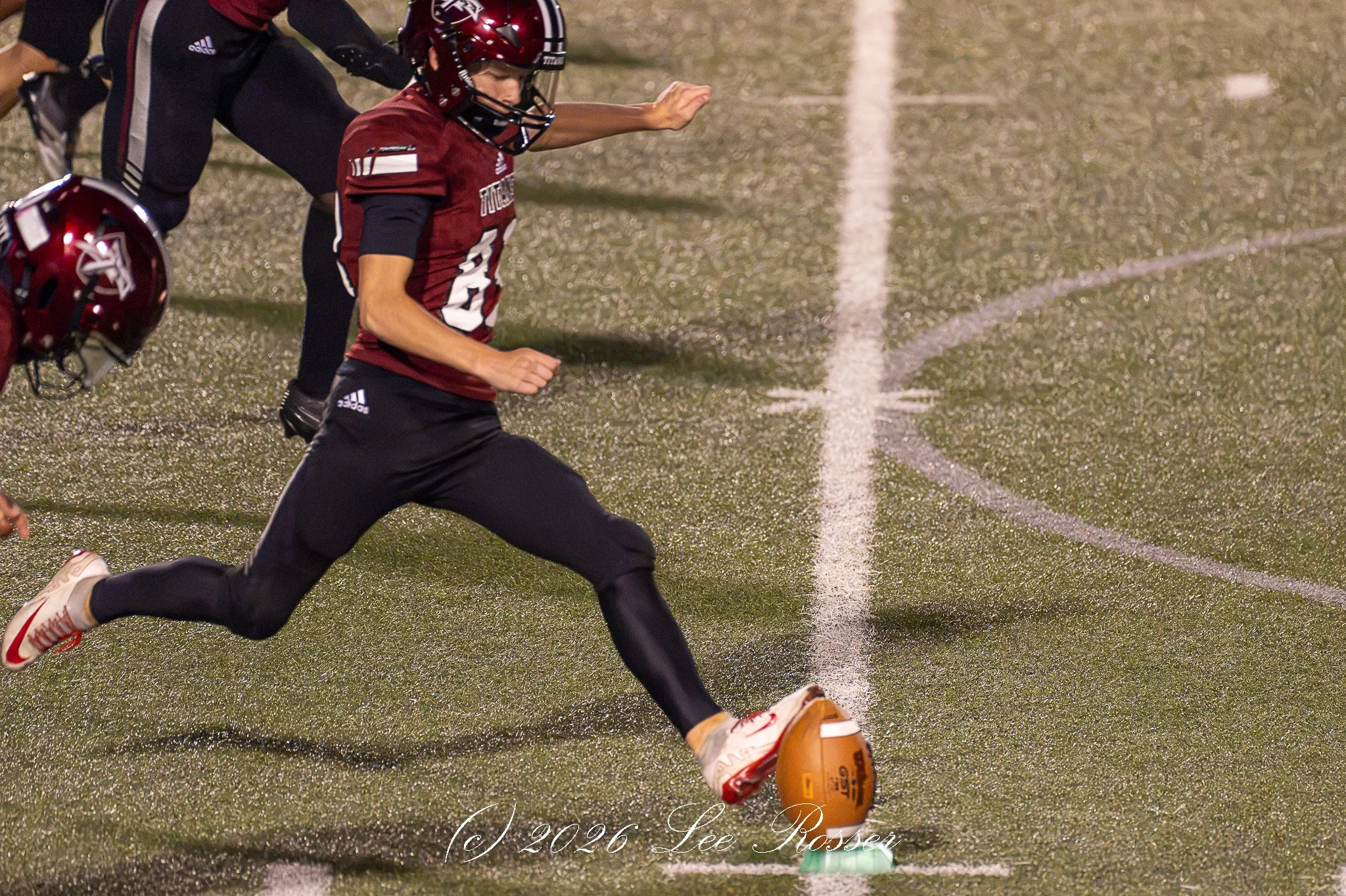 A football player wearing a red helmet and jersey is running on the field with their arm extended forward. Another teammate, also in a red helmet, is partially visible nearby. The players are on a green, textured turf football field with white lines.