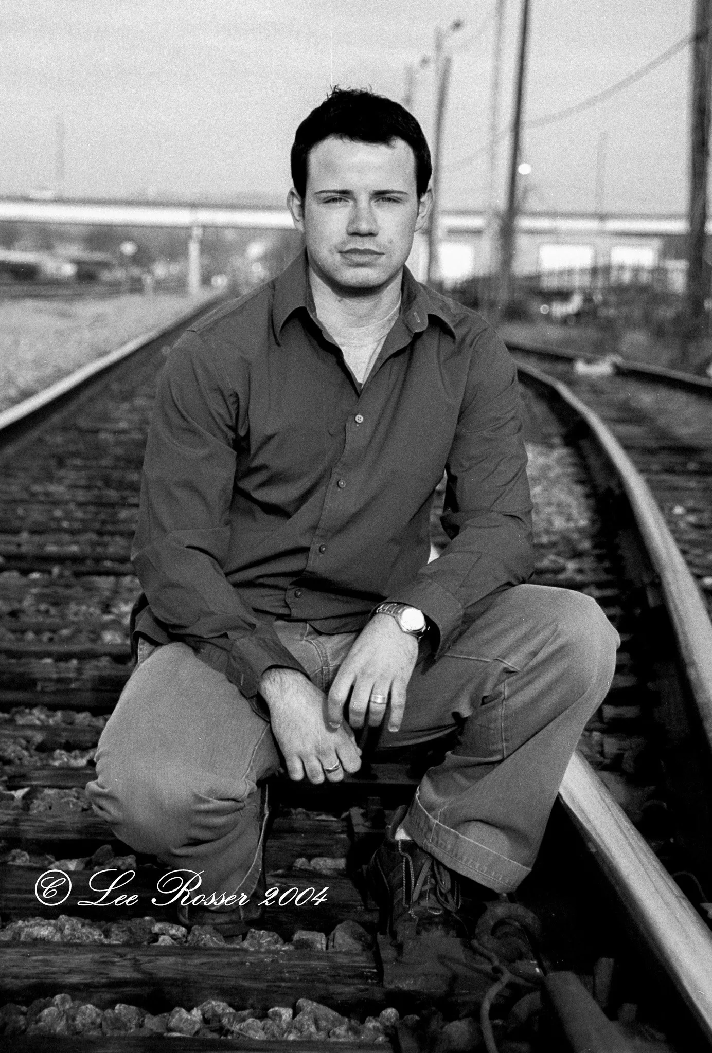 A black and white photo of a young man sitting on railroad tracks, wearing a button-up shirt, pants, boots, and a watch, with a serious expression, mountains and utility poles in the background.