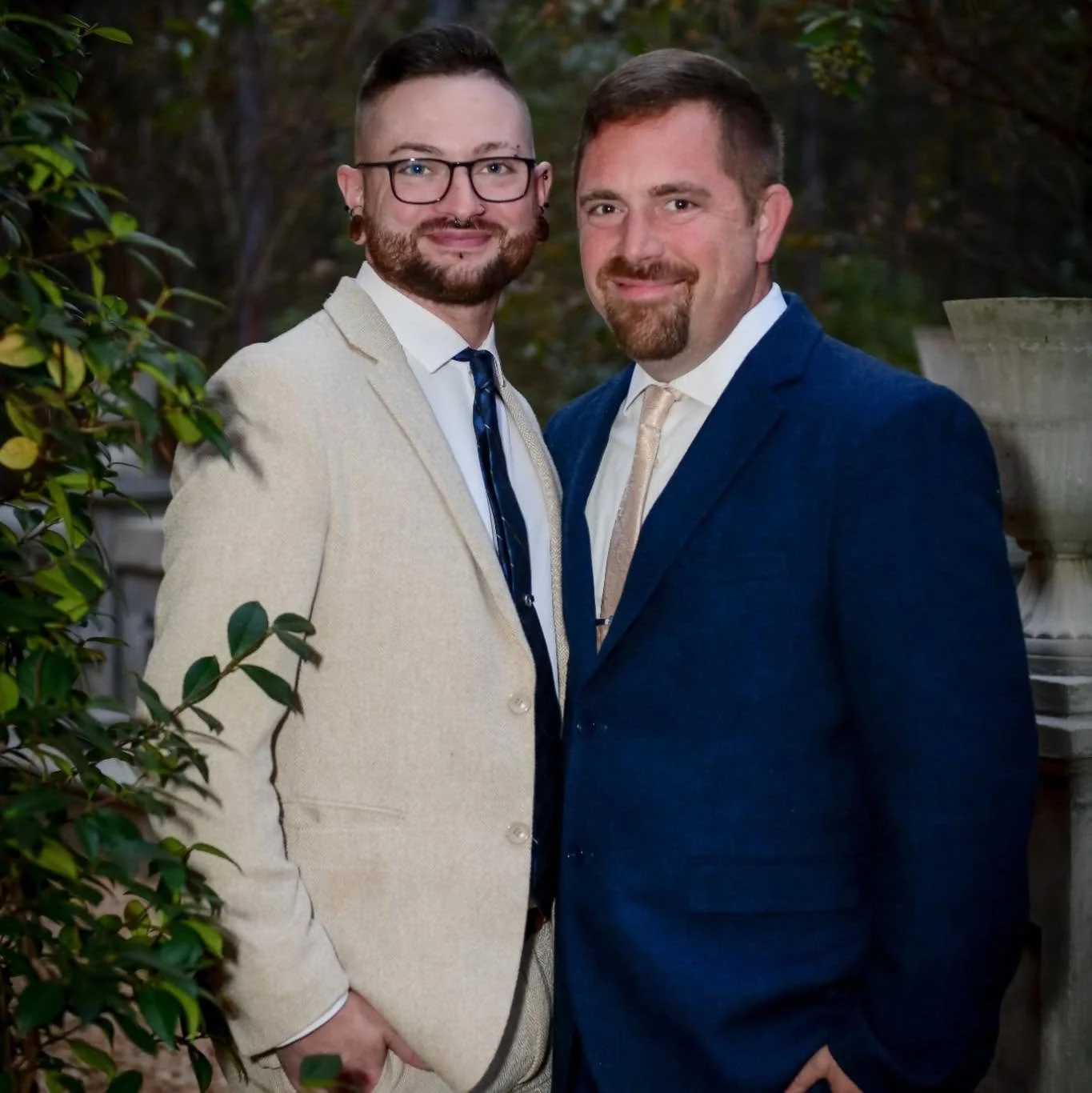 Two men dressed in suits standing outdoors, smiling at the camera, with greenery and a large plant pot in the background.