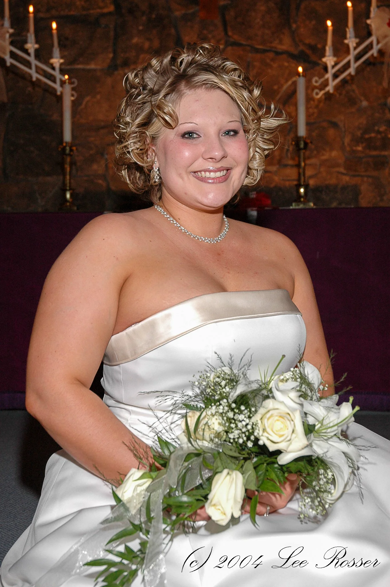 A woman in a wedding dress smiling, holding a bouquet of white roses and greenery, with candles in the background.