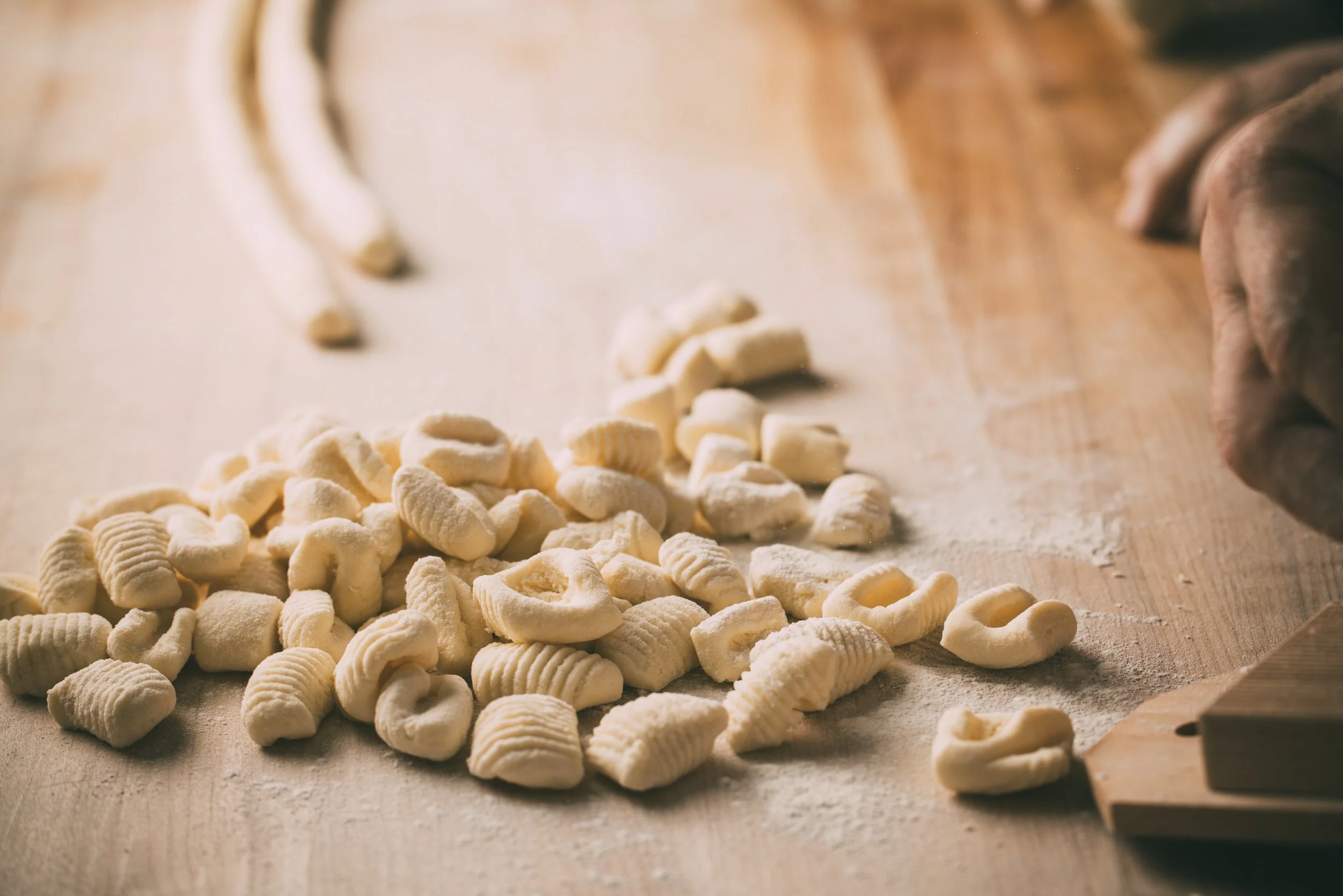 Close-up of small, ridged, white pasta pieces scattered on a wooden surface, with a hand in the background about to pick some up.