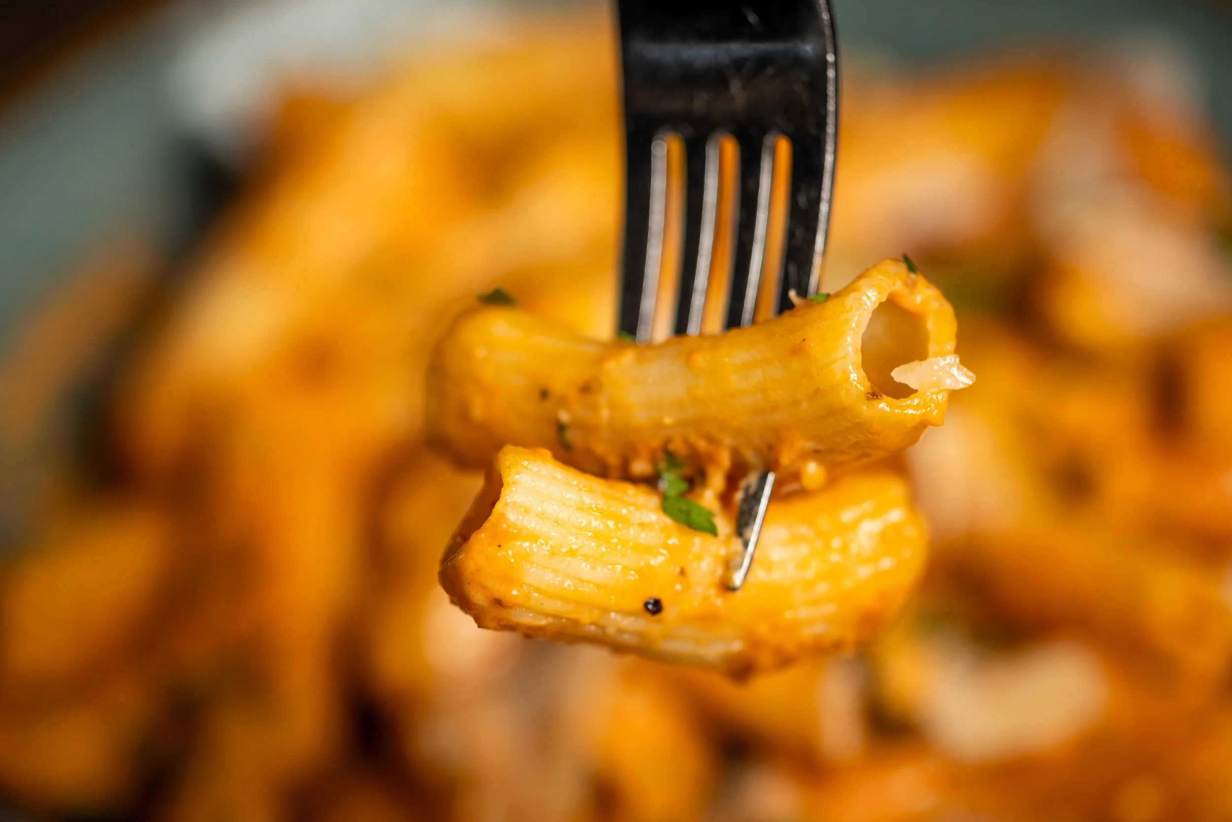 Close-up of cooked cavatappi pasta with cheese, garnished with herbs on a fork, with a blurred background of more pasta.