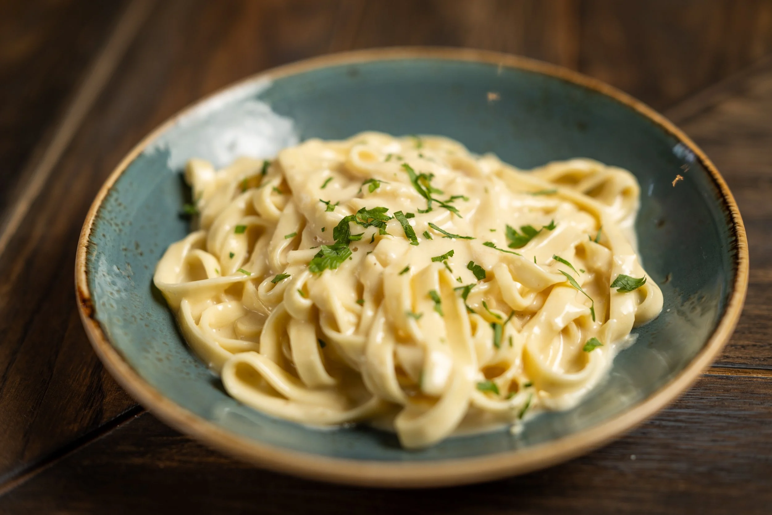 A bowl of creamy fettuccine pasta garnished with chopped parsley, served in a blue ceramic bowl on a wooden surface.