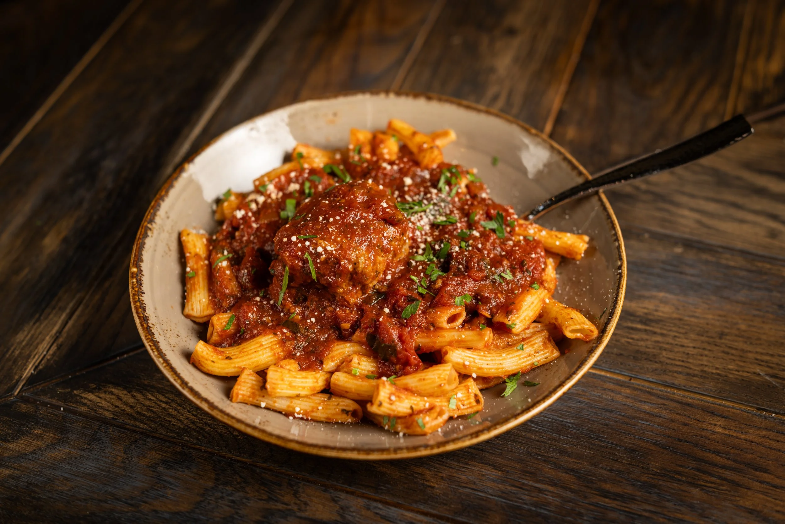 A bowl of cooked pasta with meatballs, covered in a red tomato sauce, garnished with chopped parsley and grated cheese, placed on a dark wooden table.