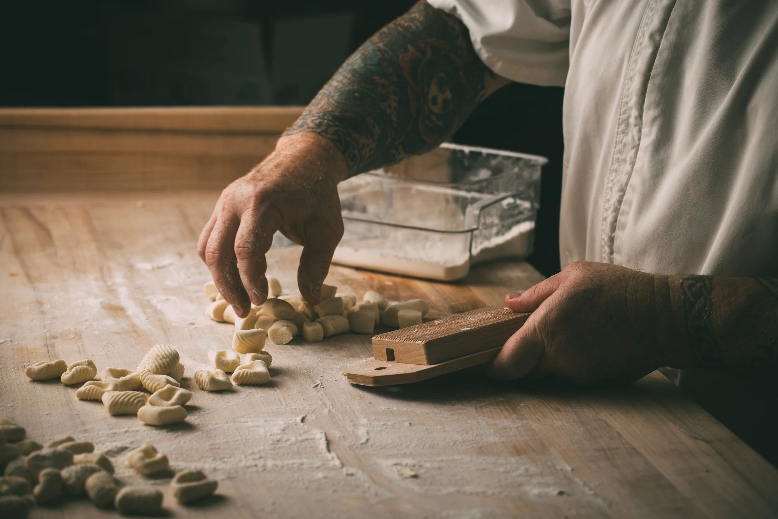 A person with tattoos on their arm, wearing a white shirt, is making pasta on a wooden surface. There are scattered small pasta pieces and a plastic container of flour in the background.