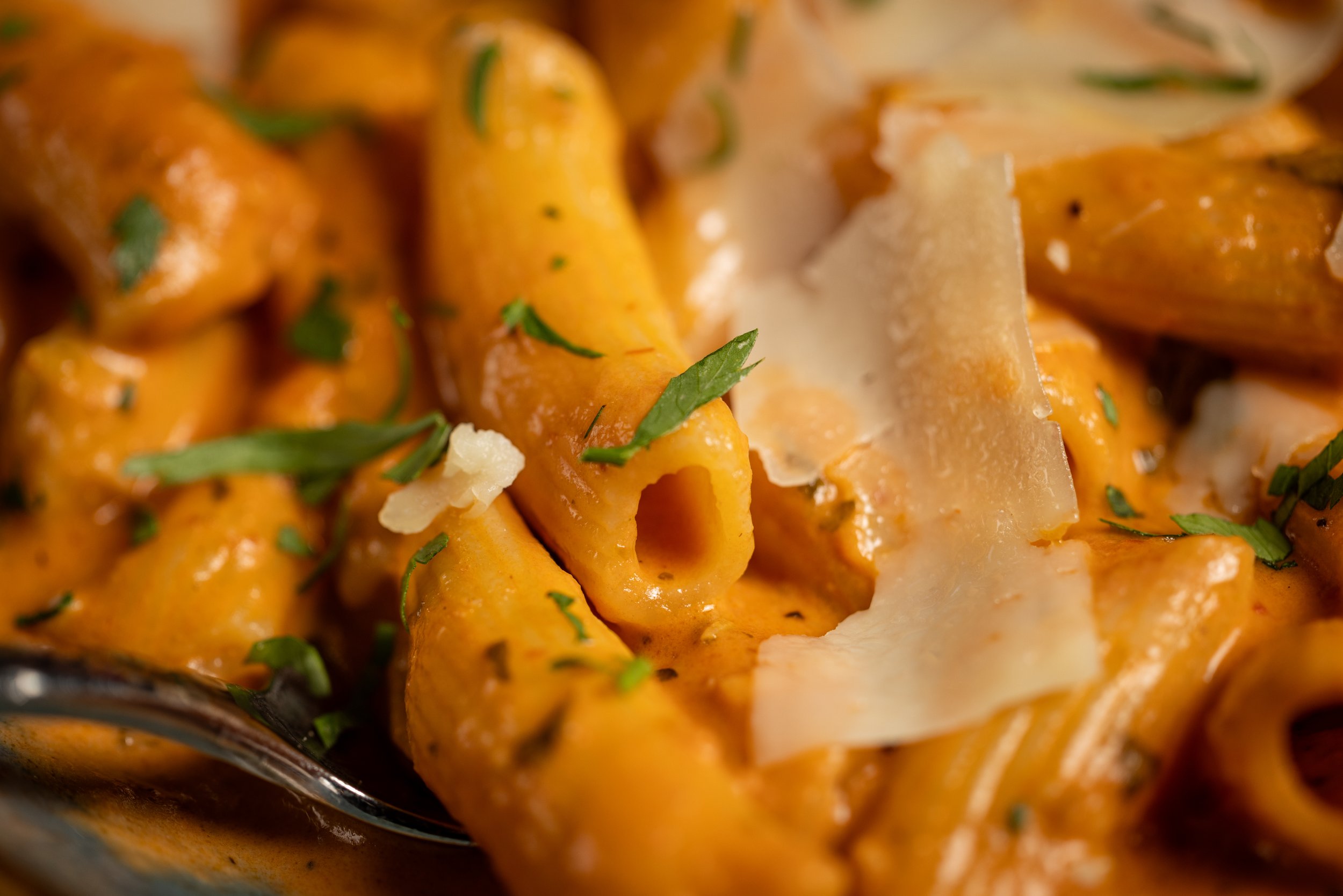 Close-up of cooked pasta with a creamy, orange-colored sauce, garnished with chopped parsley and shaved cheese.