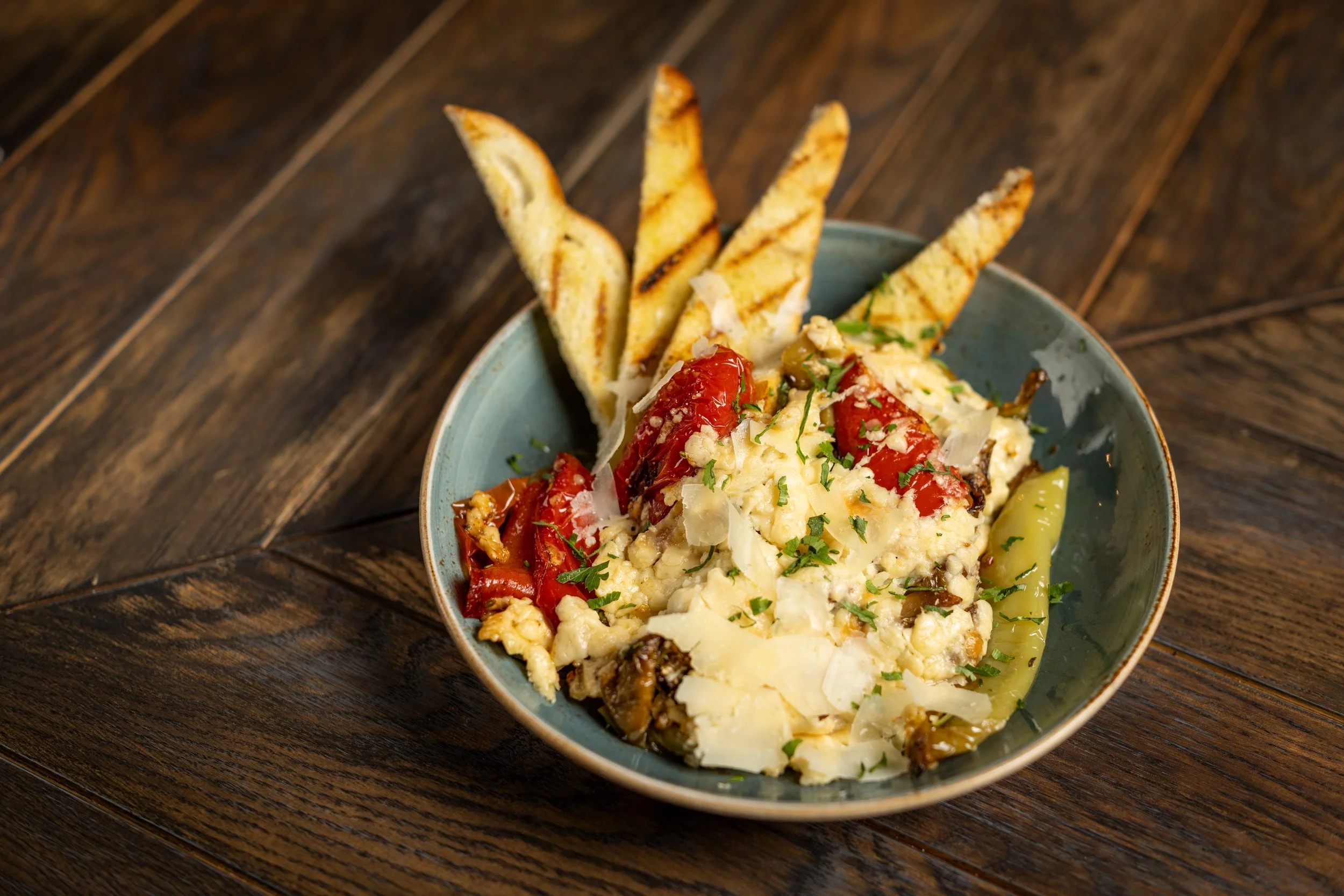 A bowl of creamy egg salad garnished with herbs and surrounded by toasted bread slices on a wooden table.