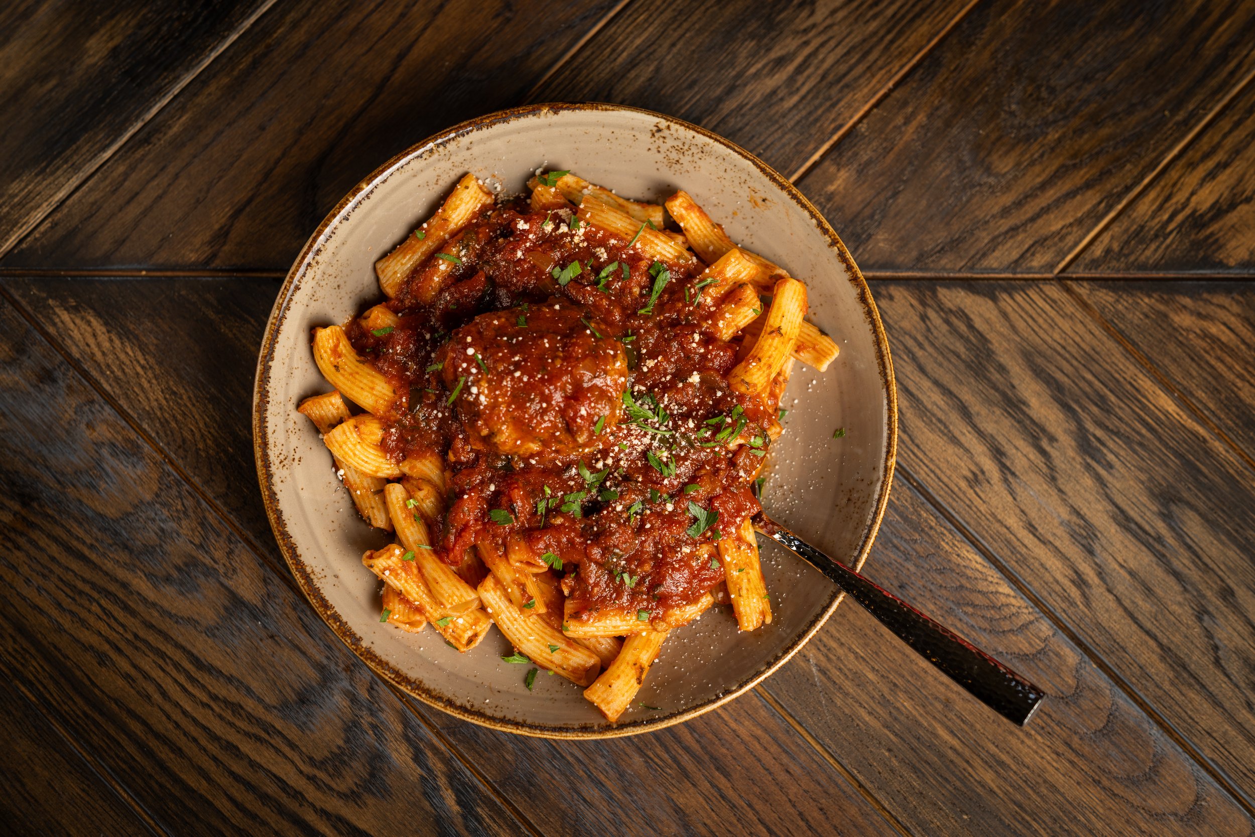 A plate of penne pasta with marinara sauce, garnished with chopped herbs and grated cheese, on a dark wooden table.
