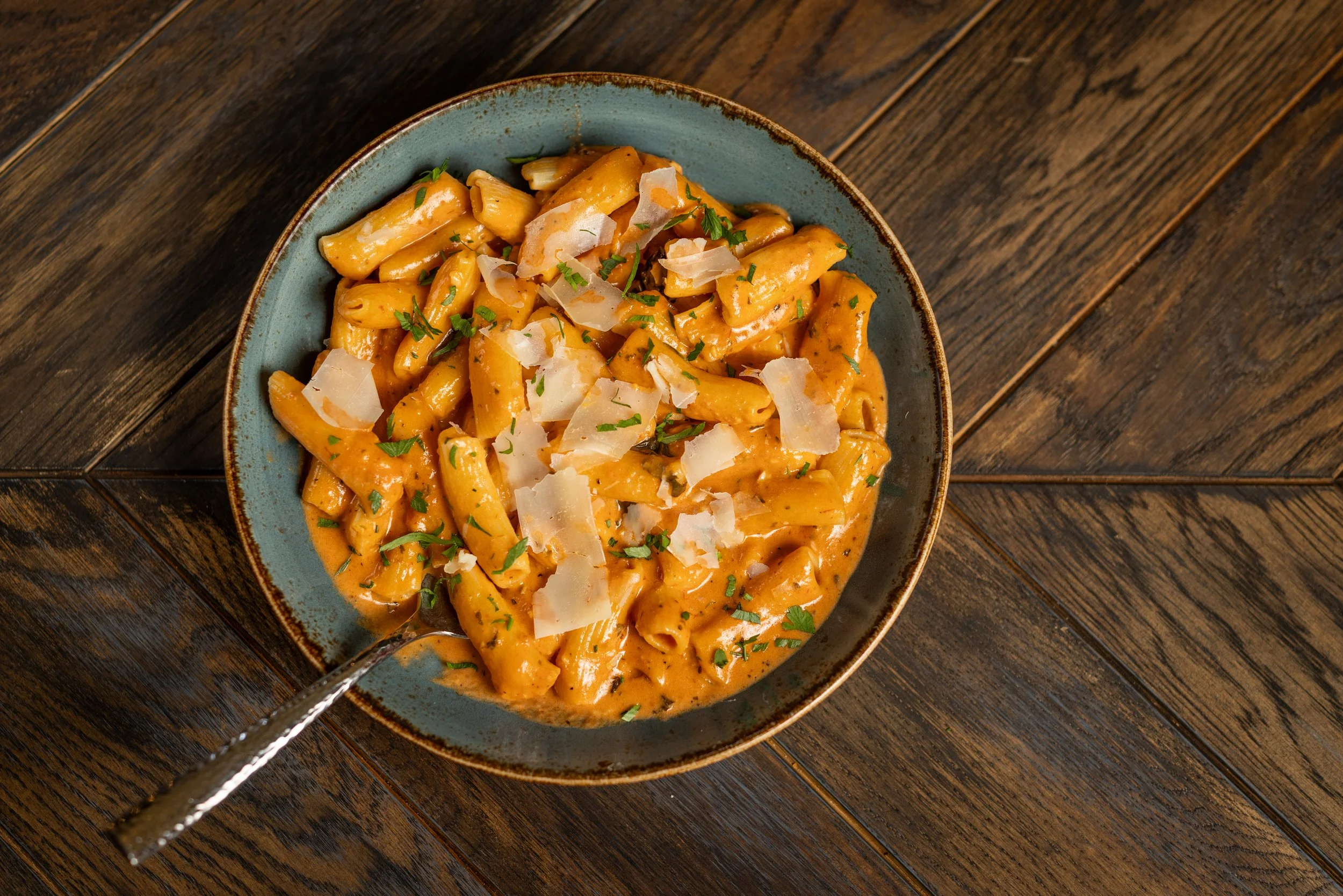 A bowl of pasta with creamy orange sauce, topped with grated cheese and chopped parsley on a wooden table.