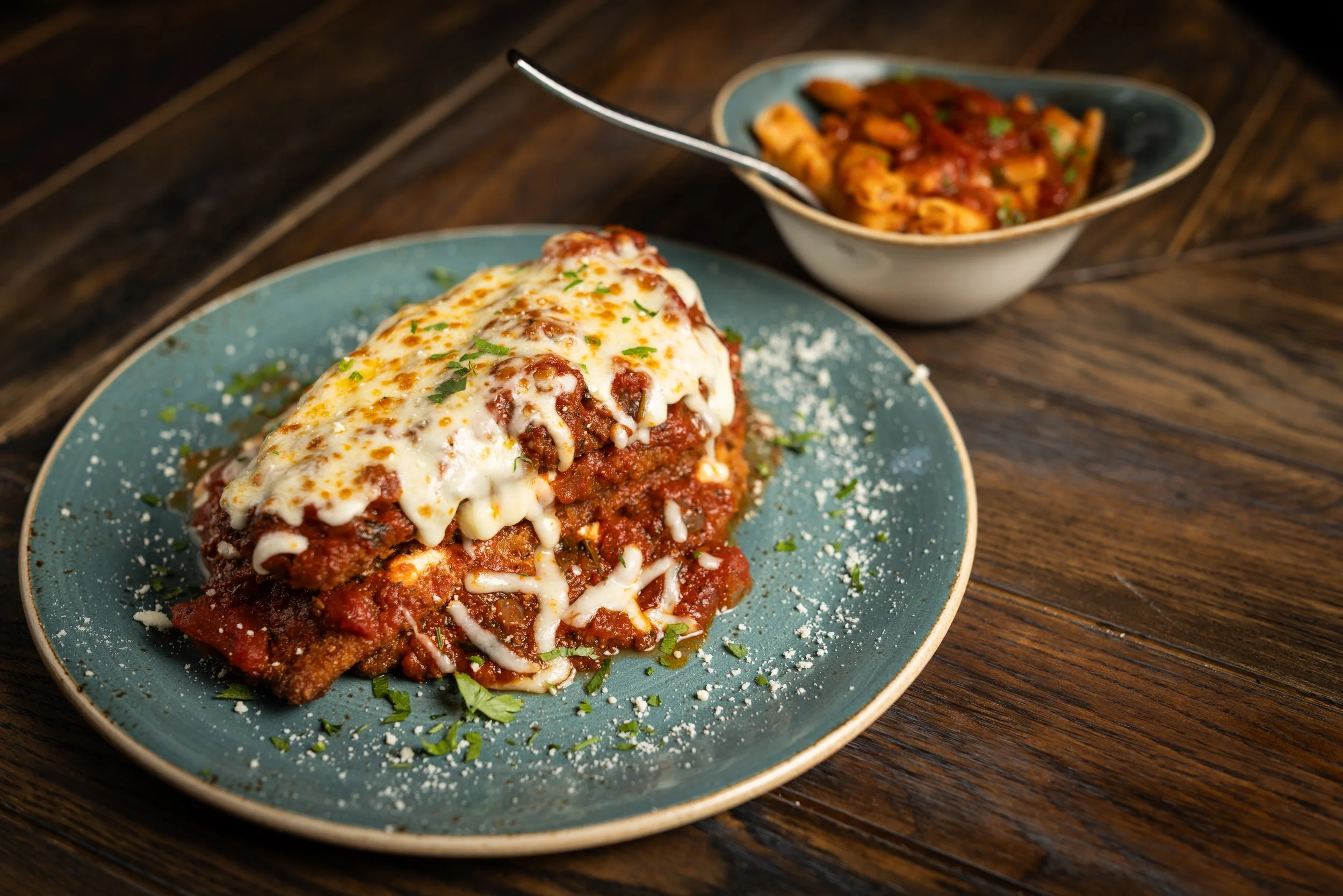 A plate of baked pasta casserole with melted cheese and tomato sauce, garnished with herbs, with a bowl of more pasta in tomato sauce in the background on a wooden table.