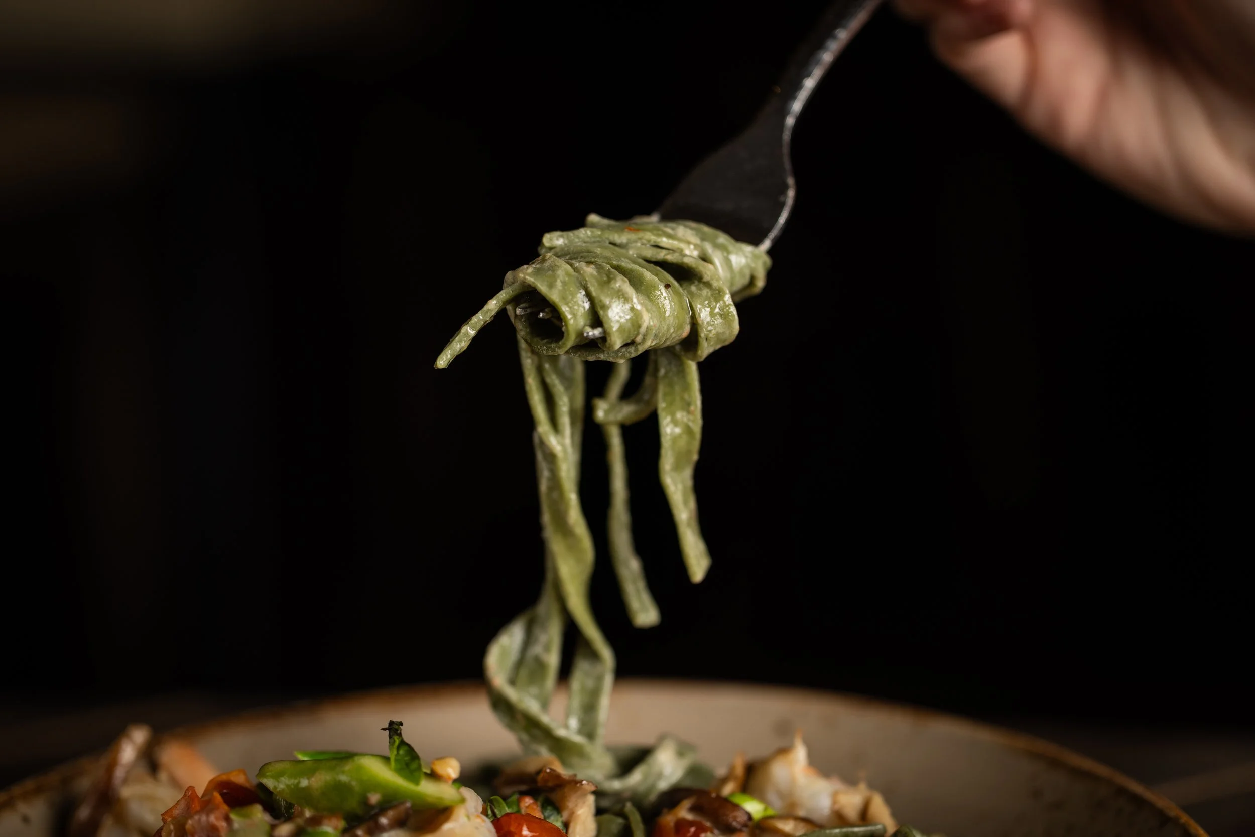 Close-up of a fork lifting a spring roll filled with green vegetables over a plate of various food items against black background.