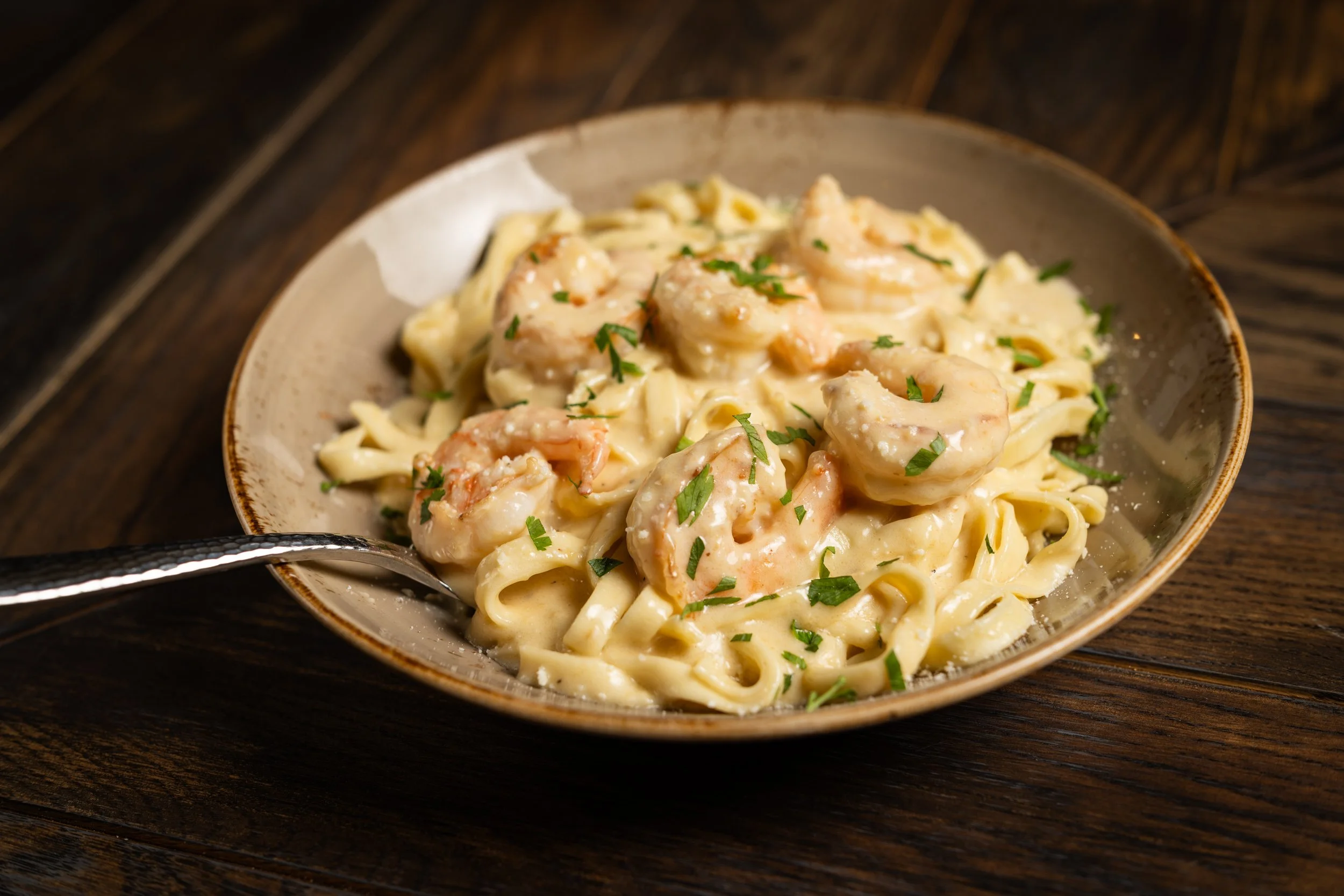 A bowl of creamy pasta with shrimp and garnished with chopped parsley on a dark wooden surface.